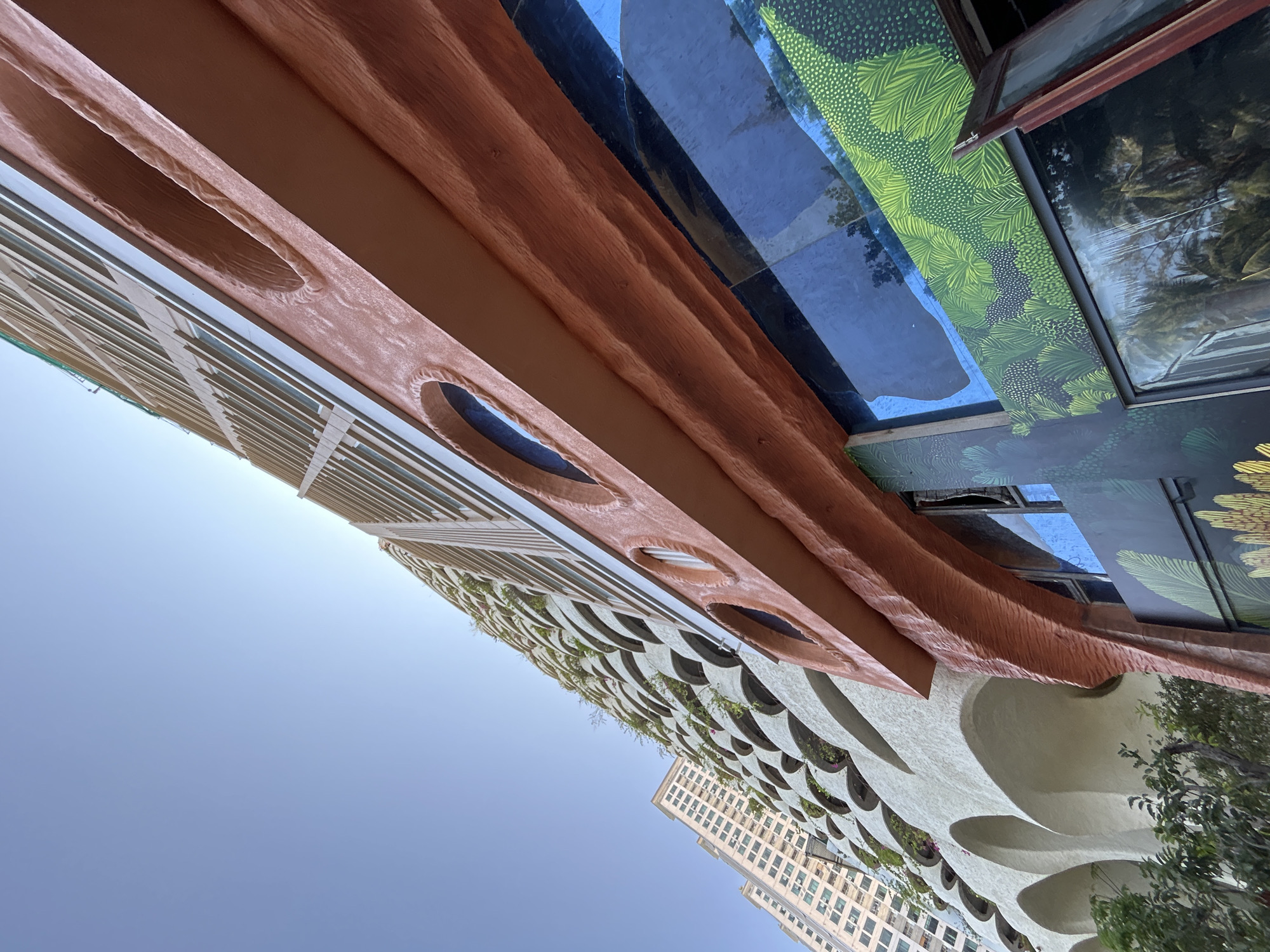 The photograph depicts an upward-angled view of buildings in what is identified as Bắc Nha Trang, Vietnam. A tall, modern structure with a facade featuring rows of circular openings containing vegetation is prominently featured. Adjacent to this building is another tall building with a more traditional, gridded facade. Above, the eaves of a building display a decorative design of concentric rings, along with a green foliage pattern on the walls above. The sky is light blue and clear. The angle of the shot suggests it was taken from street level, pointing upwards to capture the architecture.