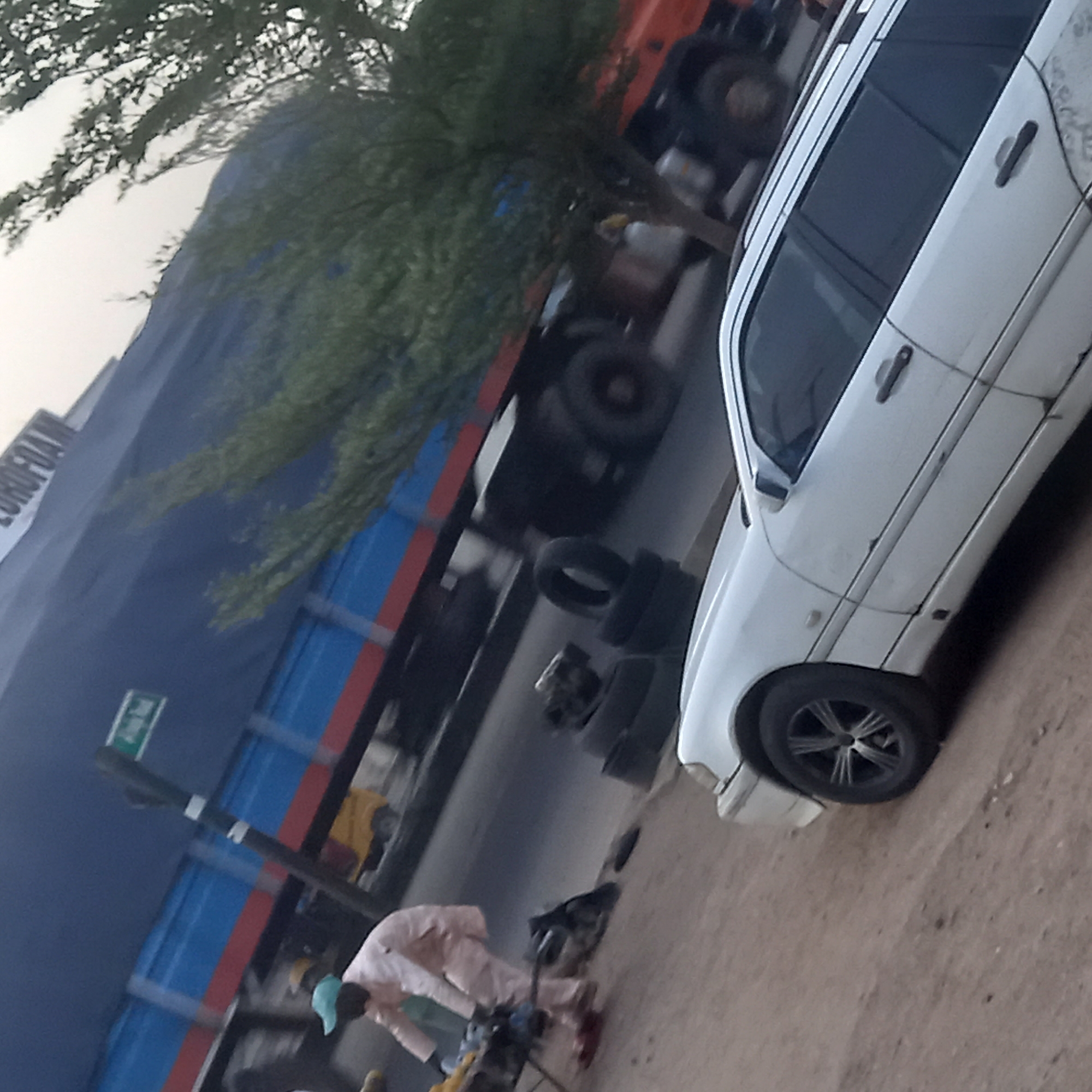 This image captures a street scene in Dadi Riba, Nigeria, featuring vehicles and a person engaged in work.
**Subjects:** The primary subjects include a large blue truck with a red stripe, a white car, and a person. The truck has a blue tarp-like covering over its cargo area, and its cab appears orange. The white car, possibly a sedan or hatchback, shows some signs of wear and tear, including minor dents and scuffs. A person, dressed in light-colored clothing and wearing a light blue cap, is seen bending down, seemingly working on something on the ground. Several loose tires are visible on the ground near the person and the vehicles. Trees with sparse foliage are present in the background and foreground, suggesting a natural element within the urban or semi-urban setting. A utility pole with a small green sign is also visible.
**Activity or event taking place:** The person appears to be performing maintenance or repair work, likely related to the vehicles or tires, given their posture and the presence of tools and spare tires nearby. The setting suggests a roadside service or an informal workshop.
**Notable details:** The time of day appears to be either late morning or early afternoon, characterized by a bright but diffused light, possibly indicating overcast conditions or a transitional period of the day. The weather seems clear and dry, with no signs of rain. The ground is dusty or sandy, typical of many roadside environments in the region. The overall scene suggests a functional, possibly commercial or industrial, area. No strong emotions are discernible from the person, who is focused on their task.
**Visible text:** On the side of the large blue truck or a nearby building, the partial text "EUROFOA" is visible. The text on the small green sign on the pole is too blurry to be read.