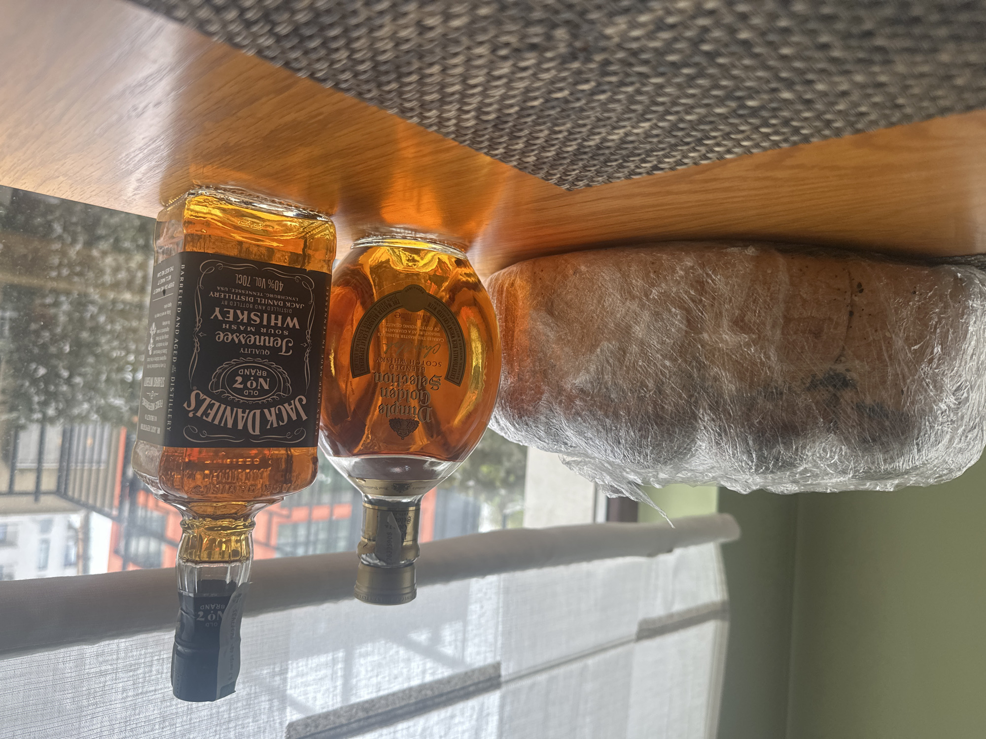 Here's a matter-of-fact description of the image:
The image shows a table or shelf with three items resting on it. From left to right, there is a bottle of Jack Daniel's Tennessee Sour Mash Whiskey, a bottle of Dimple Golden Selection Blended Scotch Whisky, and a round object covered in clear plastic wrap, potentially food. The labels on the bottles are partially visible, detailing the brand and type of liquor.
Outside the window, there is a view of a city area with buildings, and infrastructure, likely reflecting an urban setting. Given the request about the location of Sofia, Bulgaria, I can only state that these images could possibly be from Bulgaria, but cannot state if they definitively are.