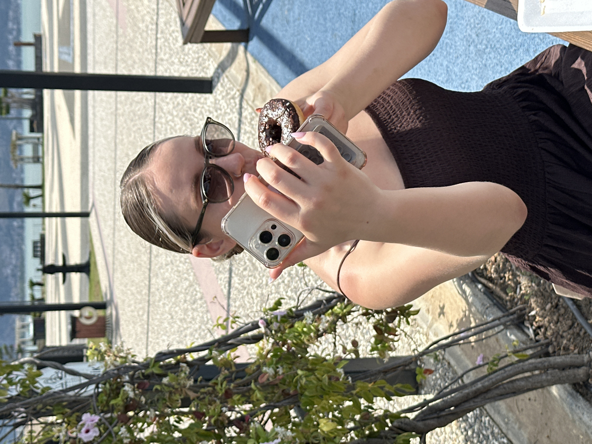 The photograph depicts a person holding a chocolate donut and two cell phones, likely taking a picture. The individual has sunglasses perched on their head, fair skin, and light brown hair with blonde highlights. They are wearing a brown, textured dress.
The setting appears to be outdoors, with a body of water and buildings visible in the background. There is a tree or shrub in the foreground with flowers and green leaves. The pavement suggests a public area.
Without additional context or landmarks, definitively pinpointing a specific location within Fethiye, Turkey, is challenging. However, based on the presence of water, buildings, and public seating, it could be along the Fethiye harbor or promenade area.