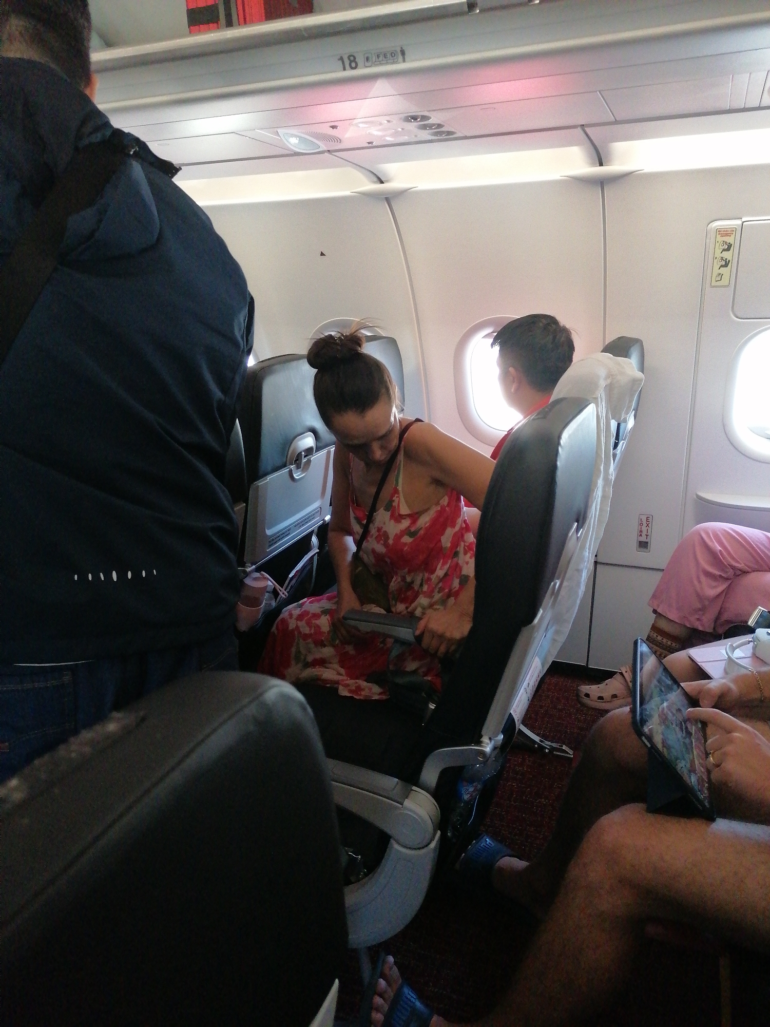 The image shows the interior of an airplane, with passengers seated. A woman with a bun is in a floral dress, seated and looking down at something in her hands. Beside her, a child is looking out the window. A person wearing a blue jacket and jeans is standing in the aisle. In the foreground, there are glimpses of other passengers, one using a tablet and another wearing pink pants. The overhead compartment shows a seat number '18'. The location is an airplane cabin.