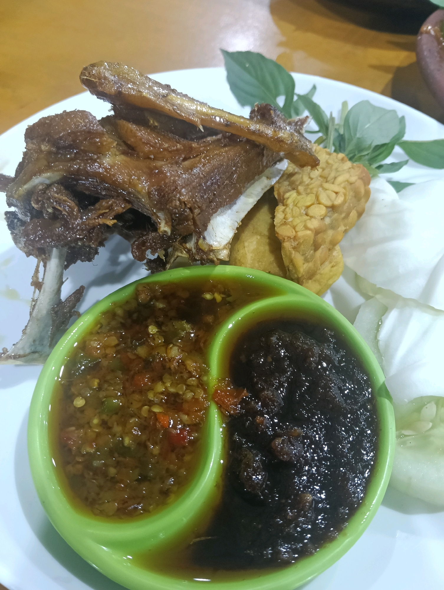 This is a close-up, high-angle shot of a plate of Indonesian food. The main subject is a piece of crispy fried duck, with its bone clearly visible. To the right of the duck are pieces of fried tempeh. In the foreground, there is a bright green, divided dipping dish containing two types of sauce: one on the left appears to be a chili-based sambal with visible chili flakes and seeds, and the one on the right is a darker, richer sauce, possibly kecap manis (sweet soy sauce) or a more complex sambal. The plate is white, and to the right of the dipping dish are slices of cucumber and some green leafy vegetables, likely for garnish. The background is blurred, showing a wooden table surface. There is no indication of a specific location within Ardirejo, Indonesia, beyond the general context of Indonesian cuisine.