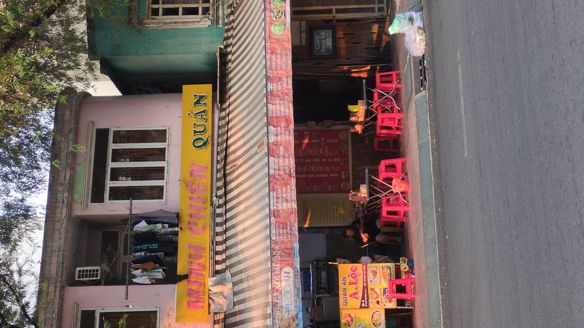 The image shows a street scene in Thủ Đức, Vietnam, focused on the front of a small eatery. A pink, two-story building houses "Quán Minh Chiến," indicated by a vertical yellow sign. A striped awning partially covers the storefront, revealing a display banner with menu items.
Inside, red plastic chairs and tables are arranged for dining. A person sits inside.
The restaurant also advertises "Quan An A. Loc" with a smaller sign displaying food images and contact information. The street is visible on the right side of the frame. A plastic bag sits on the edge of the sidewalk.
