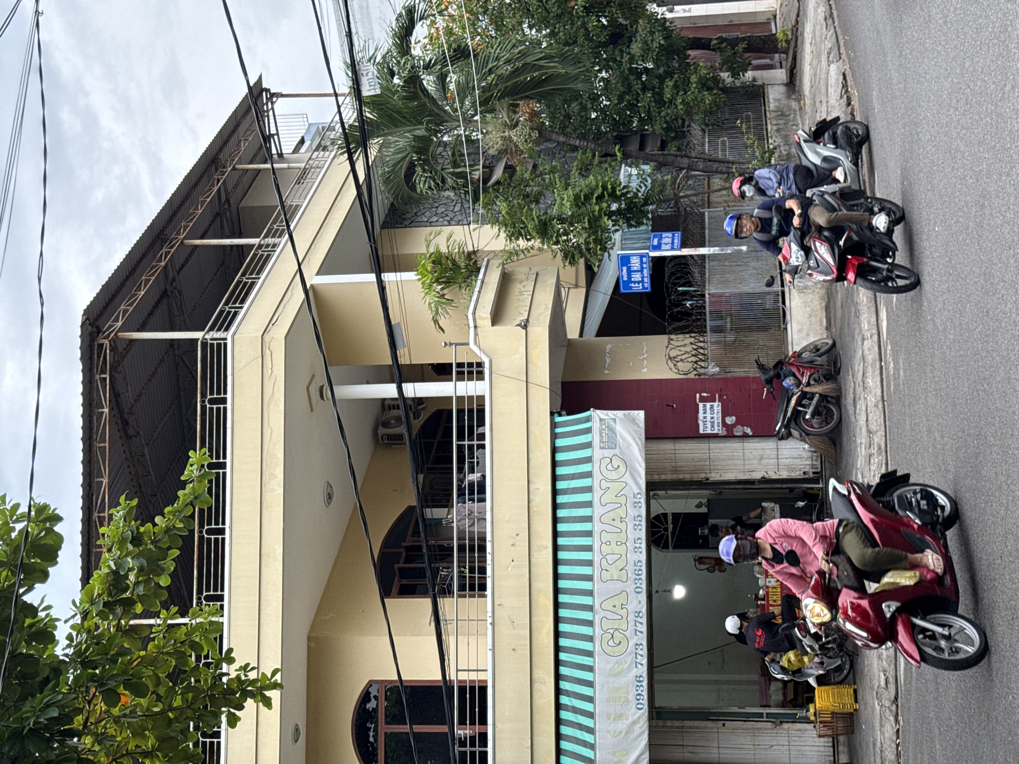 The image depicts a street scene in Nha Trang, Vietnam. In the foreground, a woman wearing a pink long-sleeved shirt, olive green pants, and a purple face mask is riding a red scooter. She is wearing a blue helmet and pink sandals. Behind her, a man in a blue helmet and a light blue jacket is riding a silver scooter with a passenger. He is wearing a dark shirt and brown pants. A red scooter is parked on the left side of the street. The buildings in the background are multi-story structures with yellow facades. A shop with a green and white striped awning has the words "GIA KHANG" in large lettering. There are also several motorcycles parked in front of the shop. Trees and power lines are visible throughout the scene, with a cloudy sky above.