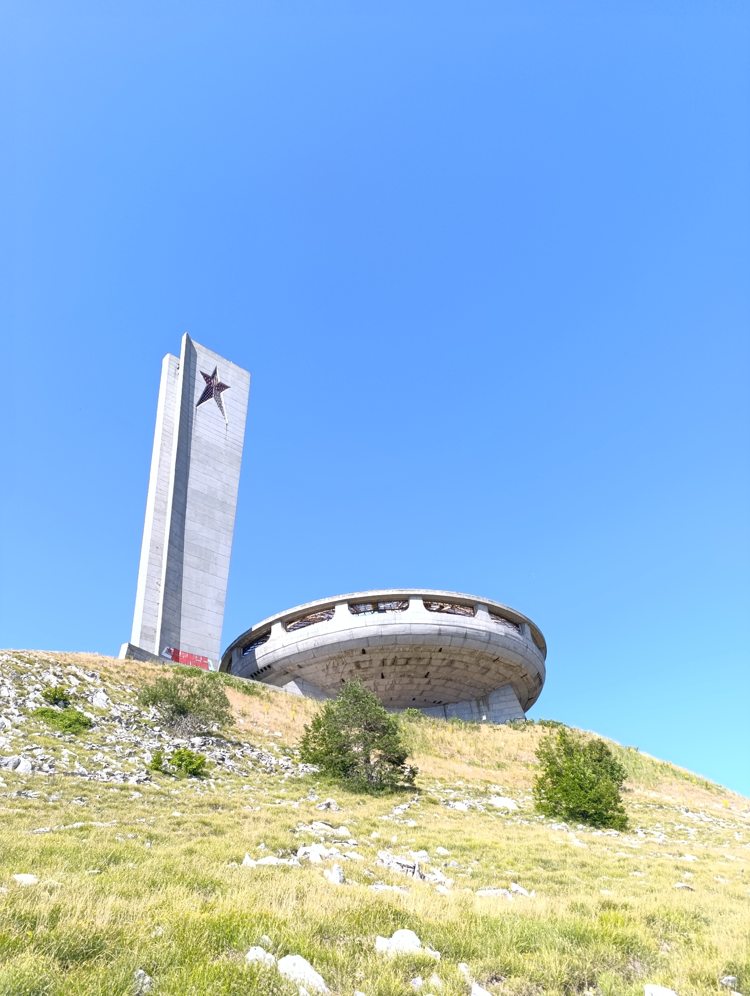 The image depicts the Buzludzha Monument, a former Communist Party headquarters situated on Buzludzha Peak near Shipka, Bulgaria. The monument complex, viewed from a grassy slope, consists of a large, saucer-shaped main building and a tall, concrete tower topped with a red star. The architecture is brutalist and modernist. The surrounding environment is a mountain landscape with sparse vegetation, including dry grass and a few small trees. The sky is clear and blue. The monument appears to be abandoned and in a state of disrepair, with some graffiti visible on the tower. There are no people or animals visible in the image.