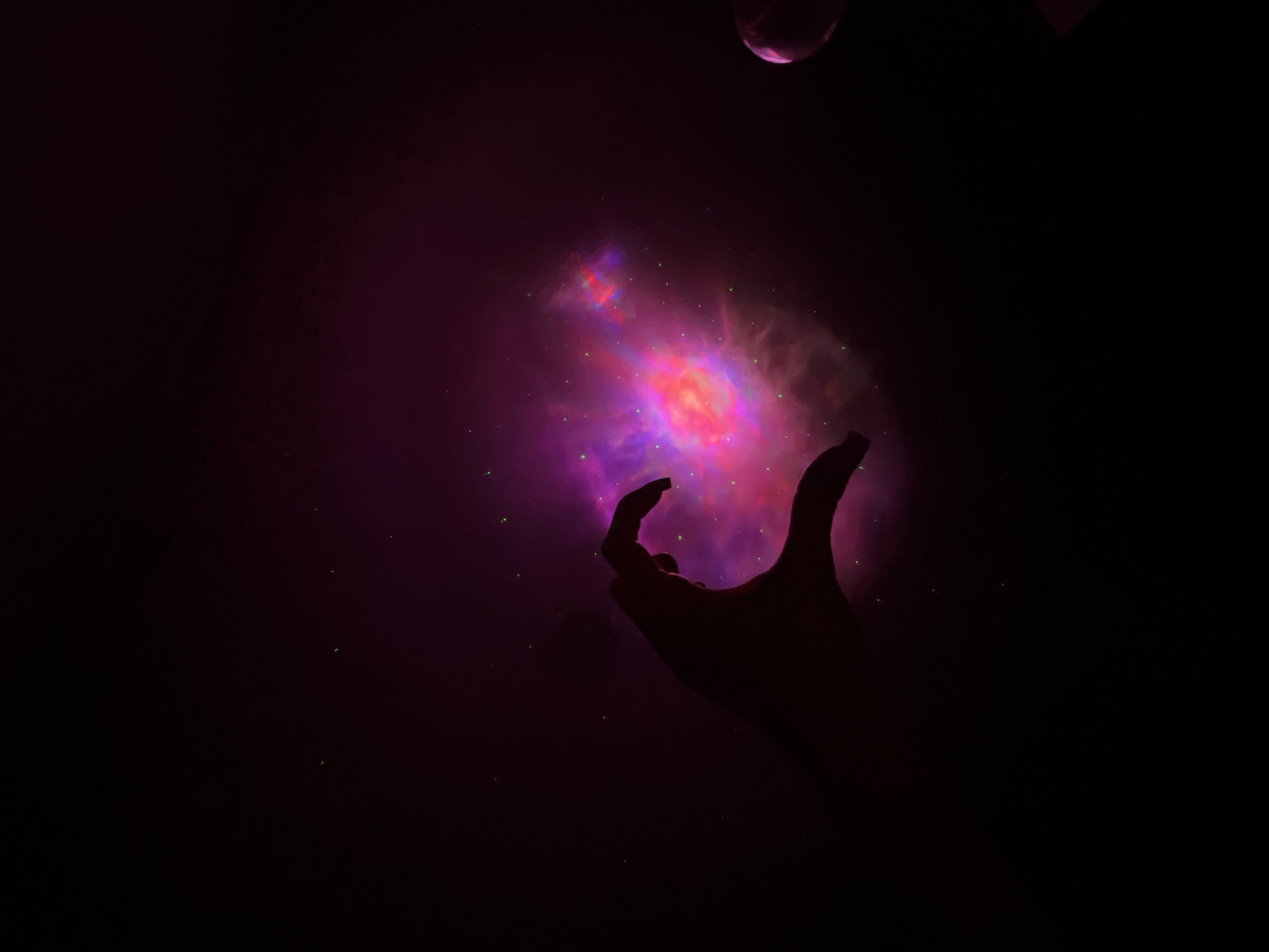 The image depicts a silhouette of a hand forming a heart shape against a backdrop of a colorful nebula. The nebula exhibits vibrant pinks and purples, with scattered green specks that resemble stars. The hand is dark and its fingers are curved to create the top curves of the heart, with the thumb and index finger meeting to form the pointed bottom. The overall lighting is dim, with the nebula being the primary light source in the center-right of the frame. To the far right, a blurry, spherical object with a pinkish hue is partially visible. The background is predominantly black, suggesting a dark, possibly nocturnal, environment. There is no discernible connection to Nizhny Novgorod, Russia in the image content.
