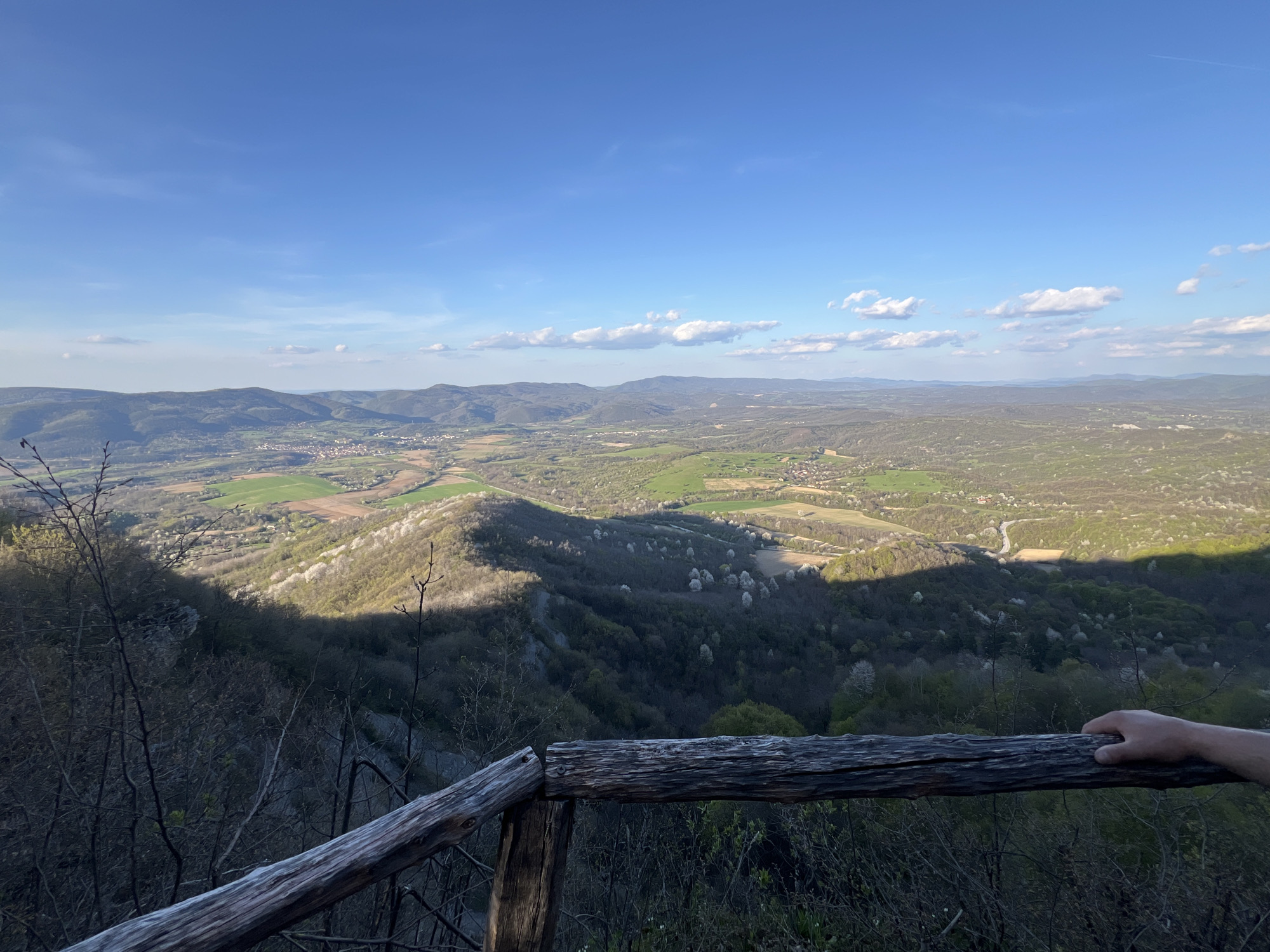 A landscape photograph taken from a high vantage point, likely in or around Elena, Bulgaria. The foreground features a weathered wooden railing, with a Caucasian human hand resting on it in the lower right corner. Beyond the railing, a steep, wooded valley stretches toward a distant landscape. The valley is divided into areas of sun and shadow, highlighting the textures of the trees and vegetation. In the mid-ground, the valley transitions into a flatter, more cultivated landscape, featuring fields, small villages, and settlements. The background consists of a series of rolling hills and low mountains under a blue sky with scattered white clouds. The time of day appears to be late afternoon or early evening, indicated by the long shadows.