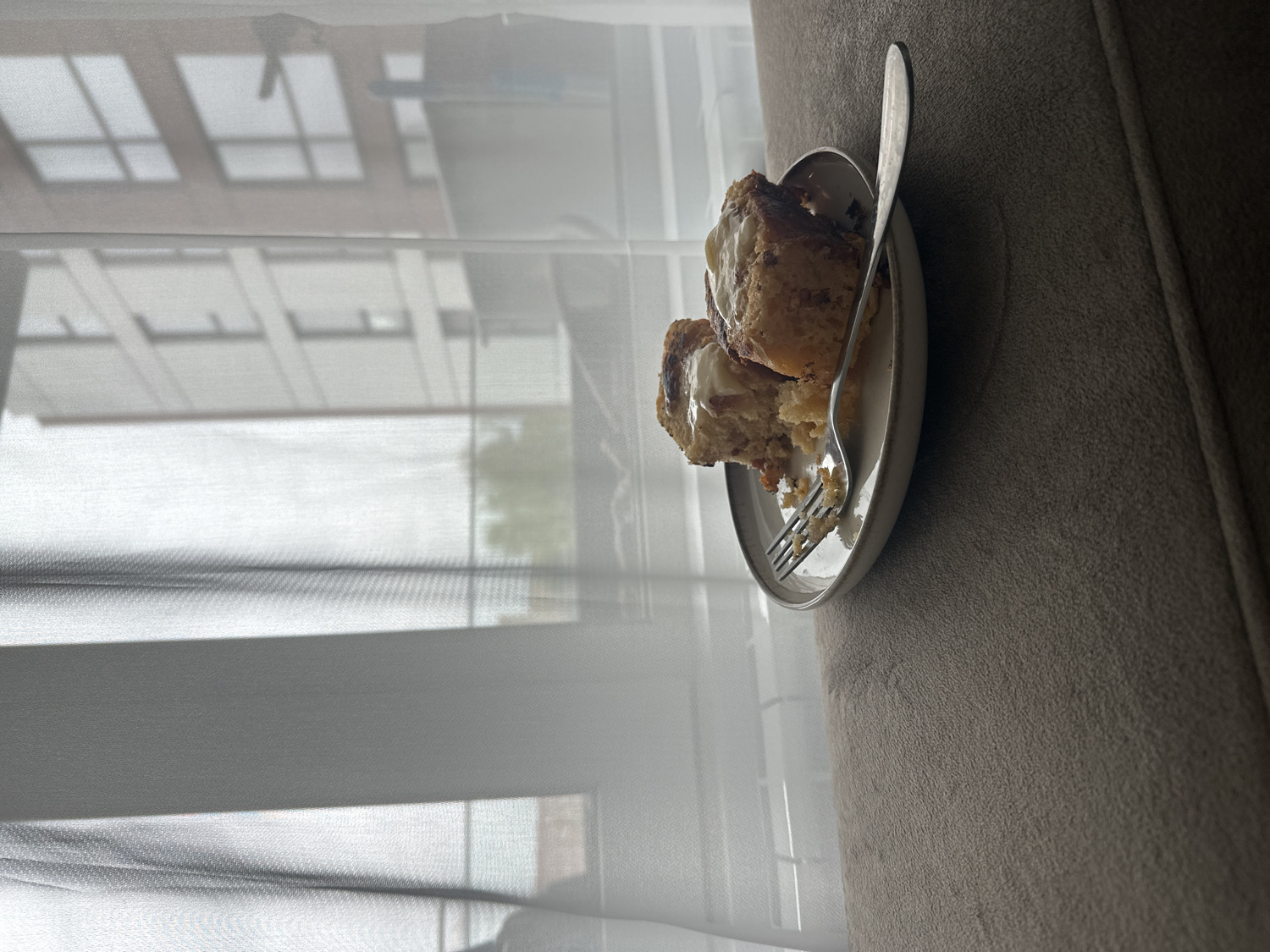 The image, rotated to a portrait orientation, depicts two pieces of a pastry, resembling cinnamon rolls or sweet buns, topped with white icing or cream cheese frosting. They are served on a small, light-colored, oval plate, where a silver fork rests with some pastry residue.
The plate is positioned on a soft, textured, light brown or beige surface, which appears to be part of a couch or armchair. In the background, to the left of the pastry, a window is visible, covered by sheer white curtains or blinds that diffuse the natural light. Through the translucent fabric, blurred outlines of external buildings can be seen, suggesting an urban or suburban environment.
The lighting is soft and natural, indicating it is daytime, possibly morning or afternoon. The scene conveys a quiet, relaxed, and intimate moment, focusing on the enjoyment of the pastry indoors. There are no people present, and no visible text. The overall atmosphere is calm and comfortable.