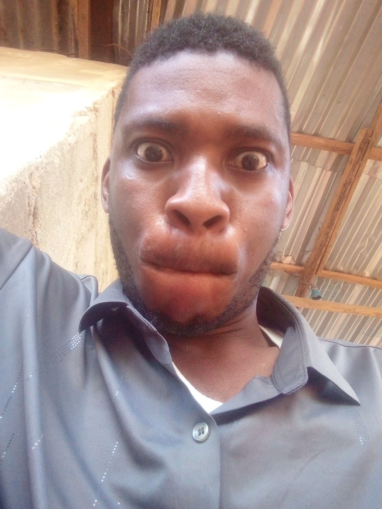 A close-up selfie shows a man with dark skin and short, dark hair. His eyes are wide open, and his lips are puckered out. He has a short beard. He wears a dark gray button-down shirt. The background consists of a concrete wall on the left and corrugated metal roofing on the right. The location is in the city of Unknown, Unknown.