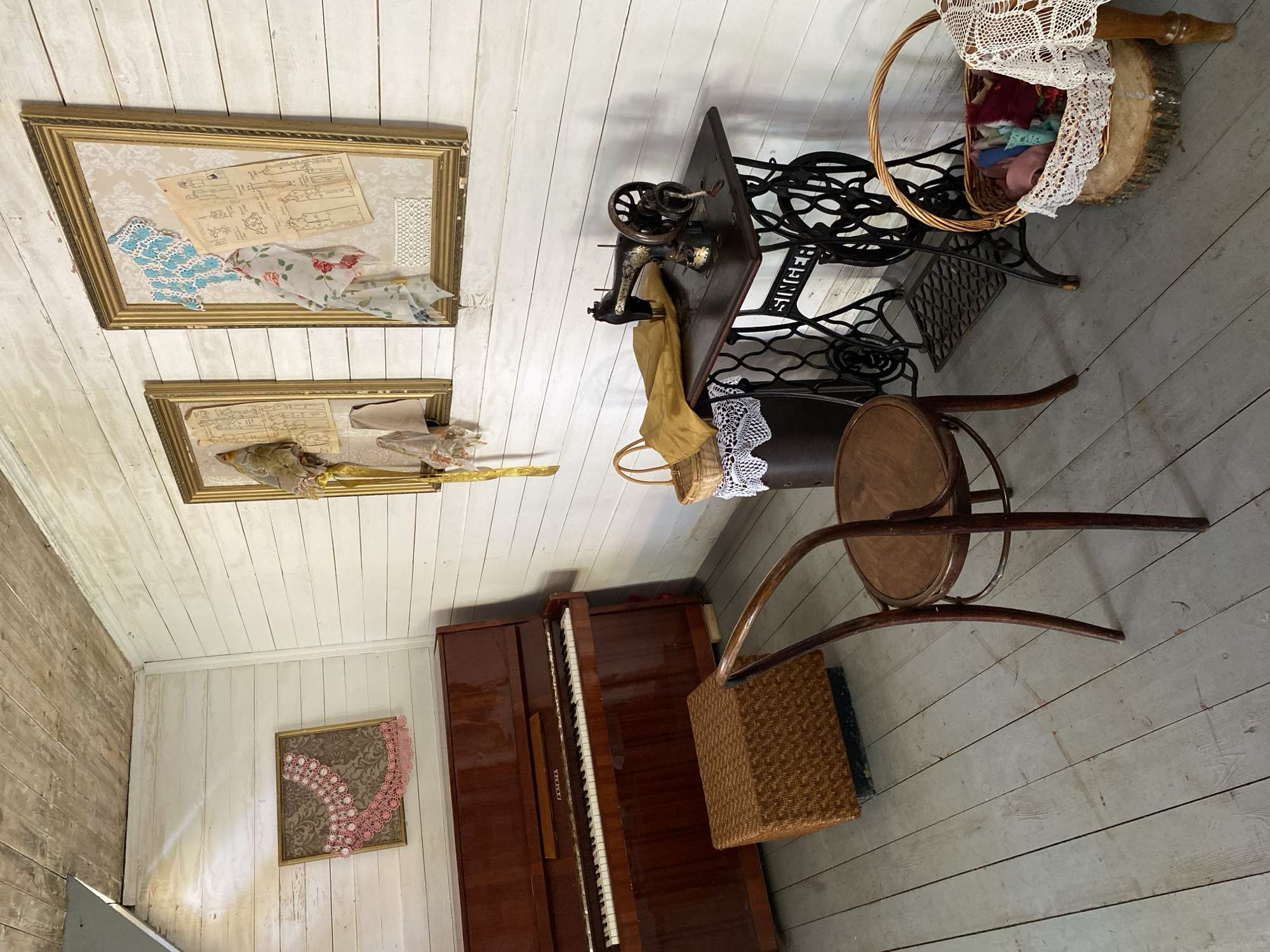 The image depicts an interior scene of a room with wooden walls and floor, suggestive of a historical or rustic setting. On the left, a polished wooden upright piano with a keyboard visible is situated next to a woven square stool. Above the piano, a framed artwork features decorative pink lace.
The right side of the room is dominated by a vintage black Singer sewing machine on a dark wooden table with ornate cast-iron legs. Beside the sewing machine, a basket holds a yellow fabric. A cylindrical dark object covered with white lace is also present. A wooden chair with a curved back is positioned in the foreground between the piano and the sewing machine.
On the wall, two framed pieces are displayed. The left frame contains several pieces of fabric and what appear to be sewing patterns. The right frame holds similar items, including a blue lace piece and more sewing patterns. A basket filled with colorful fabric scraps, resting on a wooden log, is located near the sewing machine. A portion of a table with a white lace tablecloth is visible at the bottom right. The overall ambiance is that of a creative workspace from a bygone era.
This scene is located in the Votkinsk Museum of M.I. Tchaikovsky, Votkinsk, Russia. The room is likely a representation of a creative space within the composer's former residence or a related historical building.