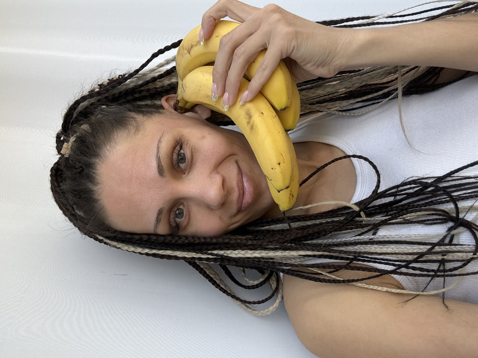 A woman with multi-colored braids lies tilted on a white surface, her head near the top left corner of the frame. She is holding a bunch of bananas near her ear with her right hand. The woman has fair skin, light eyes, and wears a white tank top. The image does not provide information about the location in the city of Rusanovo, Russia.