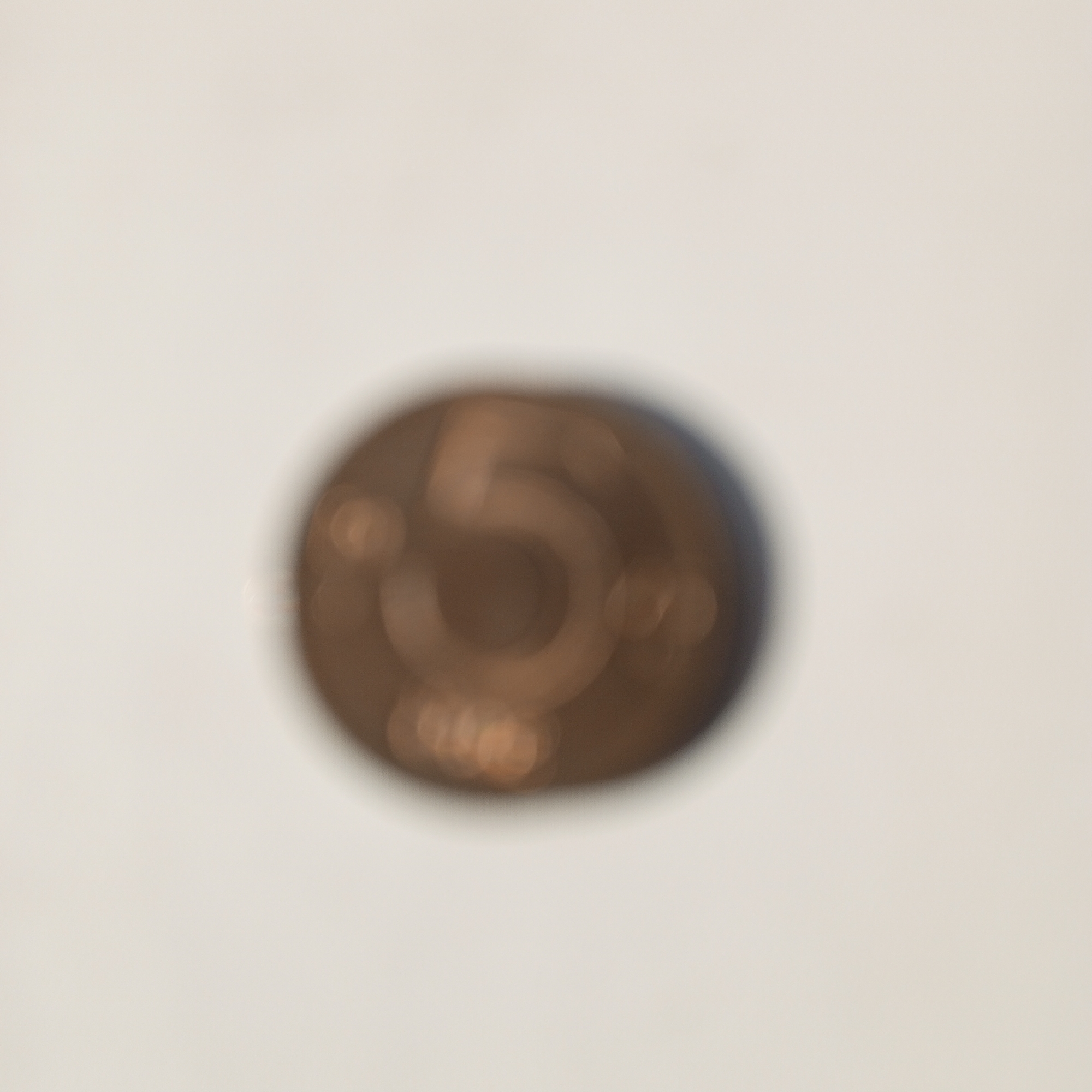 A blurred, close-up, eye-level, medium shot of a single, round, copper-colored coin is centered against a light-colored, out-of-focus background. The coin has a large numeral "5" prominently displayed in the center. The lighting appears to be even, with no strong shadows. The image is a photograph of an object, not a scene in a city. No location in Sayanogorsk, Russia, or any other location, is depicted or implied.