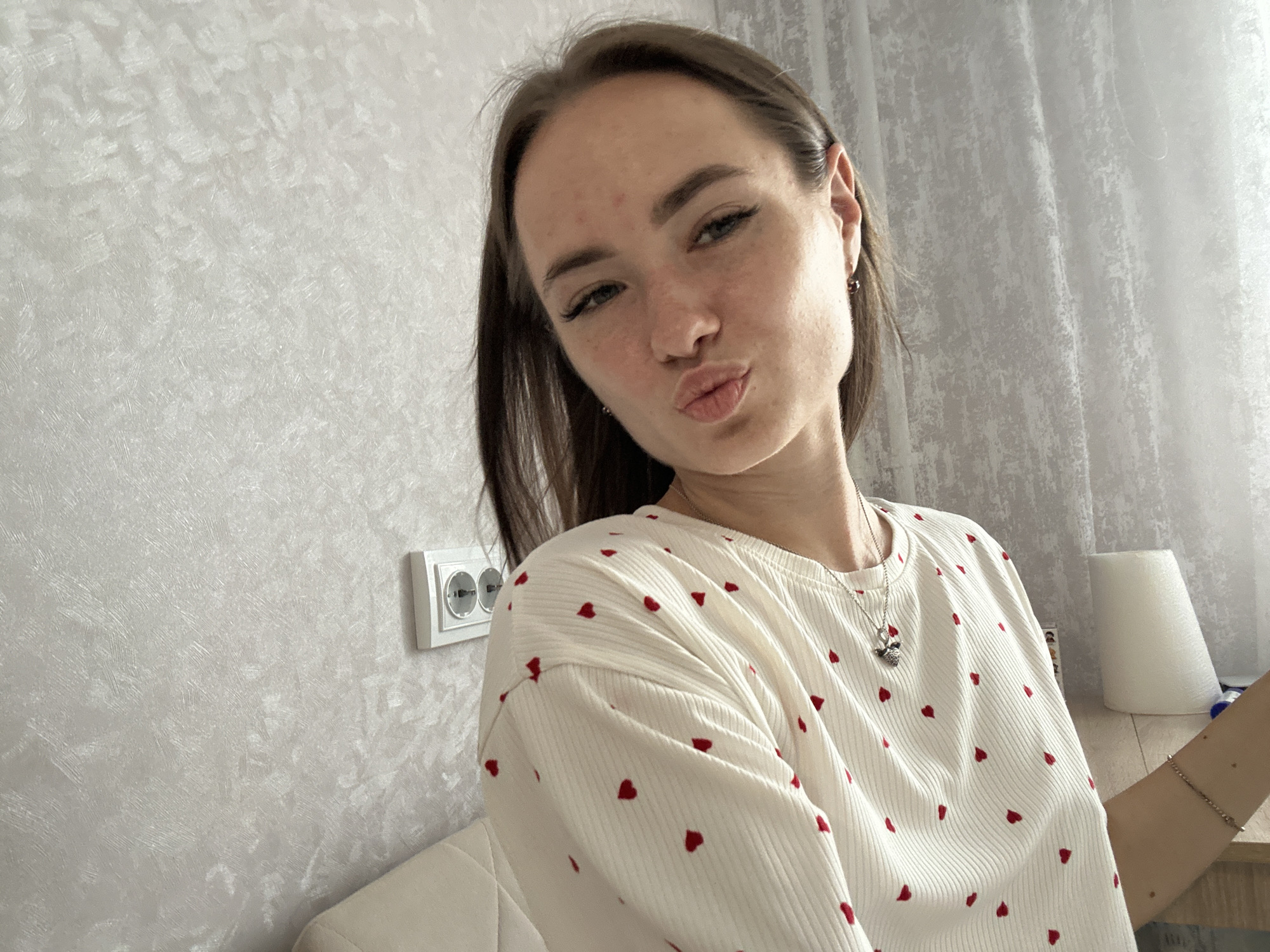 A young woman with dark hair is taking a selfie. She is wearing a white shirt with red hearts printed on it and a silver necklace. She is puckering her lips and looking at the camera. The background is a light-colored wall with a textured pattern. There is also a white electrical outlet on the wall. The image is taken indoors.