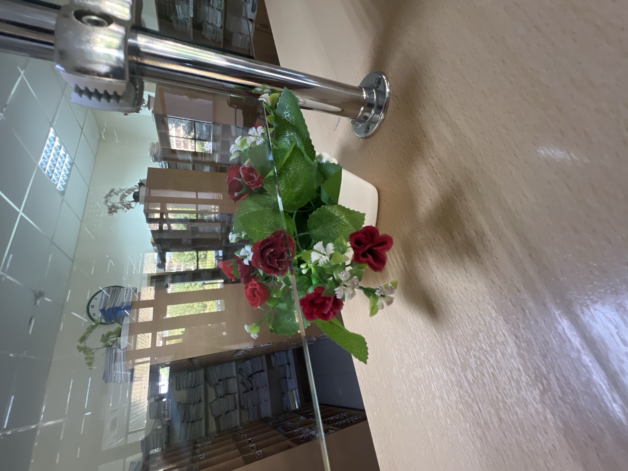 The image depicts an indoor scene, likely a library or office space, with a focus on a small bouquet of artificial red roses and white flowers in a white pot on a light-colored wooden counter. Behind the counter, a reflective surface, possibly glass, offers a distorted view of the environment. Visible through the reflection are shelves filled with books and papers, a circular clock on the wall, fluorescent ceiling lights, and what appears to be an office chair. A chrome-finished fixture with a serrated edge is prominently positioned to the right of the flowers. The overall impression is one of a tidy, well-lit interior. There is no visible indication of the city of Magas, Russia, or any other specific location in the image itself.