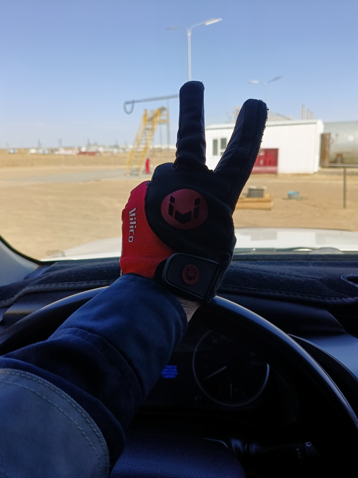 A first-person perspective shot shows a gloved hand giving a peace sign. The hand is wearing a red and black glove with a logo. The scene appears to be taken from inside a vehicle, with a steering wheel and dashboard visible. Outside the vehicle, there is an industrial or construction site with buildings and scaffolding. The sky is clear and blue. The location is specified as Unknown, Kazakhstan.