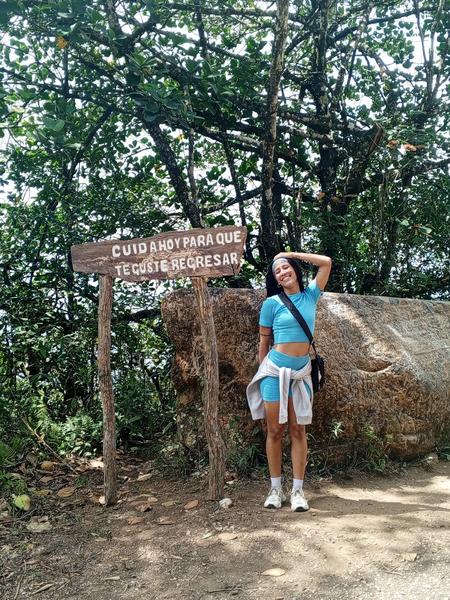 A young woman stands in a natural, outdoor setting, likely a park or hiking trail. She is wearing a light blue crop top and matching shorts, a white sweatshirt tied around her waist, white sneakers with white socks, and a blue headband. A black cross-body bag hangs from her shoulder. She smiles and has her right arm raised with her hand on her head.
Behind her is a large, light brown rock, and to her left, a wooden sign with the Spanish inscription "CUIDA HOY PARA QUE TE GUSTE REGRESAR" (Take care today so you'll like to return). The sign is attached to two wooden posts. The background is filled with lush green trees and foliage, suggesting a wooded area. The ground is a mix of dirt, leaves, and small rocks. The location is consistent with a natural area, likely near Las Piedras, Venezuela, based on the content of the sign.