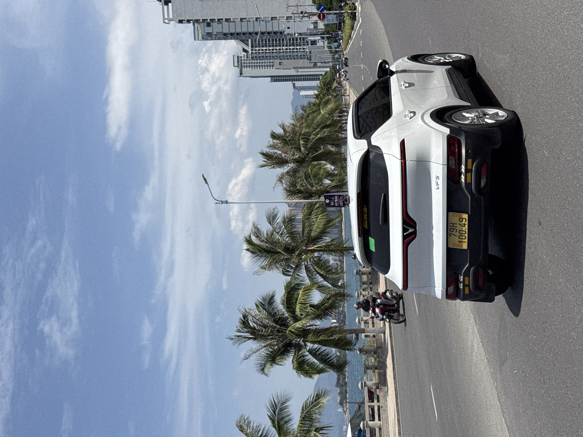 The image shows a white car with a Vietnamese license plate "79H 100.49" driving on a road in Nha Trang, Vietnam. The car is in the foreground, with its rear clearly visible. To the left of the road, there is a body of water, likely the sea, with a stone barrier and palm trees lining the coast. In the background, modern high-rise buildings are visible, indicating an urban environment. A person on a motorcycle is also seen in the distance, traveling in the same direction as the car. The sky is blue with some clouds, suggesting a sunny day. The scene appears to be a coastal road in Nha Trang, likely along Tran Phu Street, known for its beachfront hotels and resorts.