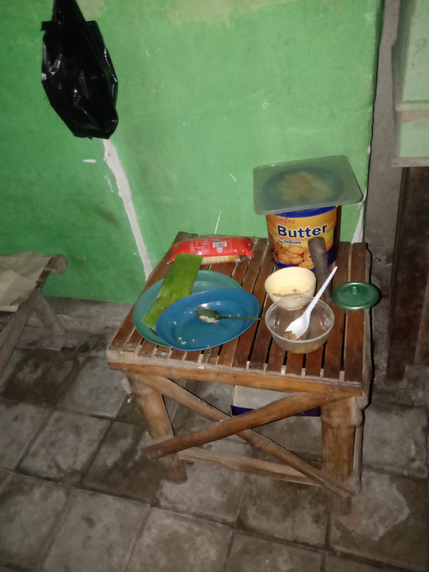 In an indoor environment with green walls and a tiled floor, a wooden table is set with various items. A black plastic bag hangs on the wall. On the table, a large blue and yellow tin labeled "Butter Cookies" sits next to a wooden mortar and pestle. Two plates, one blue and one green, are stacked on the table, with a green leaf placed on top of the blue plate, which also contains some white crumbs. A white plastic spoon rests inside a clear plastic bowl that contains some liquid. Another white bowl is partially visible behind the plastic bowl. To the right of the table, a small green lid is placed on the floor. The scene appears to be a domestic setting within the city of Jumput, Indonesia.