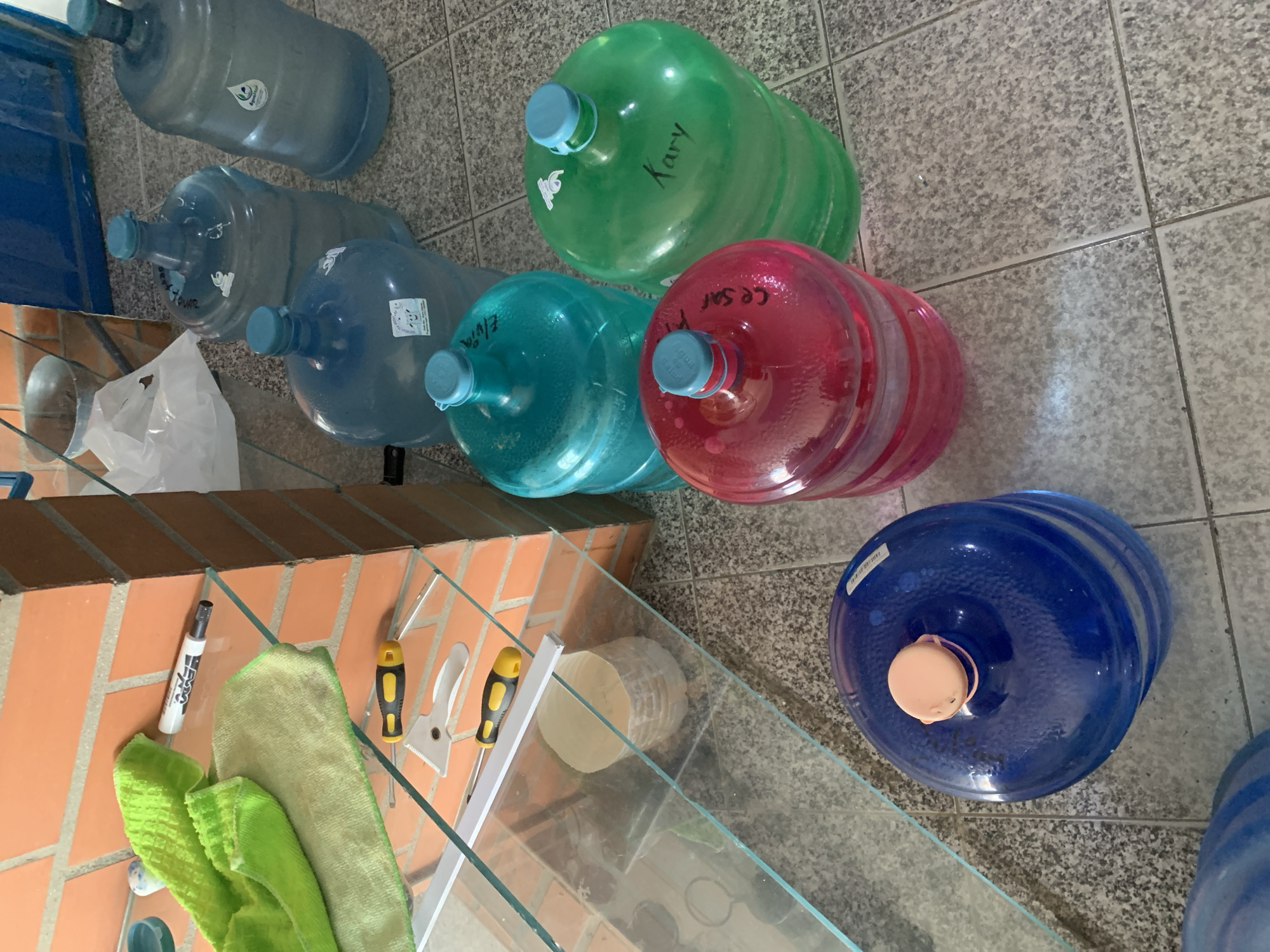 The image captures an indoor scene, likely a utility or storage area, characterized by a collection of large plastic water bottles (garrafons) arranged on a tiled floor. The floor features light gray and white speckled tiles.
Six multi-gallon water bottles are prominently displayed on the floor. From the upper left moving across and down, they include: a clear or light blue garrafon with a water drop logo sticker; another clear garrafon with the name "TOVAR" visible; a vibrant green garrafon with "Kary" written in black marker; a light blue garrafon inscribed with "Elvia" and a date, "10/7/2?"; a red garrafon bearing the name "CE SAR" (likely "Cesar"); and a dark blue garrafon with a peach-colored cap and a white barcode sticker. Other clear garrafons are partially visible on the far left.
In the foreground, a clear glass surface, possibly a shelf or tabletop, stretches across the frame. Below and behind this glass is a brick-patterned surface. Resting on or near this glass surface are several items: a white EXPO marker, a light green microfiber cloth, two screwdrivers with yellow and black handles, and a white putty knife or scraper. A white plastic bag is also visible near the brickwork, and a light-colored, cylindrical object can be seen through the glass in the mid-right foreground.
The scene suggests a domestic or small business setting in Tacarigua, Venezuela, where water is stored, perhaps for consumption or refilling. No human activity is depicted; the image is a still life of stored items. The lighting appears to be ambient indoor light.
