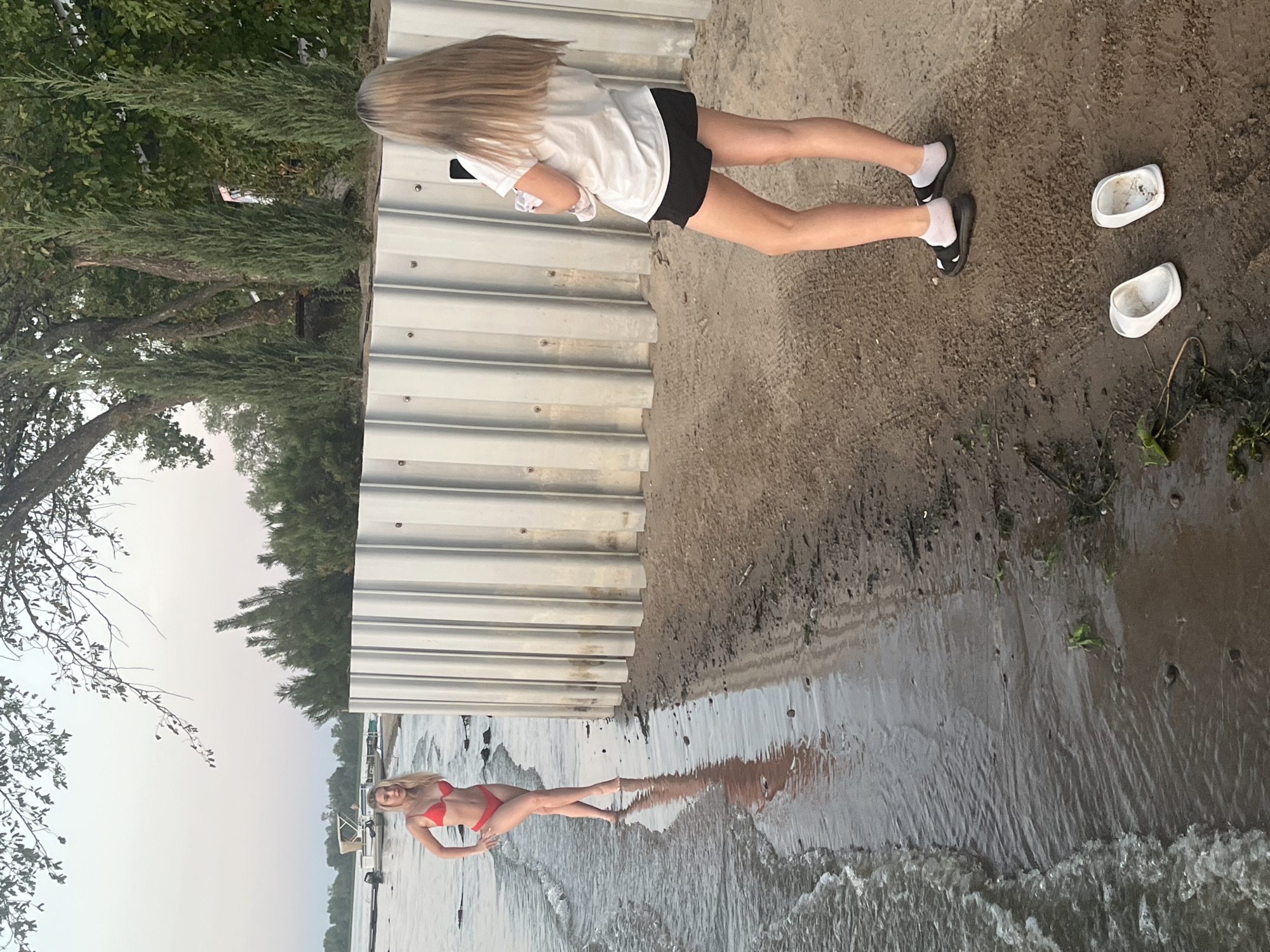 The image depicts a lakeside scene with two young women. One woman, wearing a red bikini, stands in shallow water near the shore, posing for a photograph. She has long blonde hair and her left hand is on her hip. The second woman, facing away from the camera, stands on the sandy shore and appears to be taking a photo of the first woman with a mobile phone. She has long blonde hair and is wearing a white t-shirt and black shorts. A pair of white slides are placed on the sand near her feet, and another pair of black slides with white socks are on her feet. The background features a gray metal retaining wall, trees, and a body of water with a boat visible in the distance. The overall setting appears to be a recreational area on the outskirts of Shumeika, Russia, likely during daylight hours with overcast skies.
