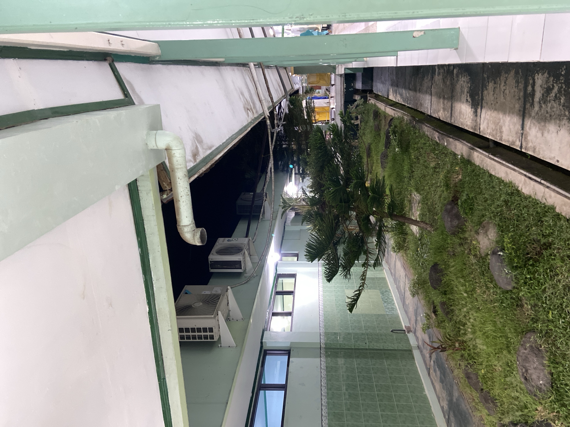 This is a landscape photograph taken outdoors, likely in Margoyoso, Indonesia, showing a view down a narrow courtyard or walkway between two buildings. The image is oriented sideways, with the top of the frame on the left.
The left side of the courtyard features the exterior wall of a building, painted a pale green or mint color. A large, white pipe curves downwards along this wall, attached to some outdoor air conditioning units mounted on white brackets. One unit is clearly visible, with a grid-like front. Above these units, the building has a white facade.
The right side of the courtyard is a strip of lush green grass, interspersed with large, dark, round stones that appear to be part of a path or decorative element. A slender tree with needle-like leaves is visible towards the top of this grassy area, casting a shadow. Further down the courtyard, on the right, is a low concrete structure bordering the grass, with what appears to be a drainage channel running alongside it. The wall of the building on this side is made of concrete panels that are weathered and stained.
Further down the courtyard, there are windows visible on the left building, with dark frames and reflections suggesting indoor lighting. The courtyard itself appears to be an outdoor space between buildings, possibly part of a residential complex, school, or other institutional facility. The lighting suggests it might be late afternoon or early evening, with ambient light illuminating the scene. There are no people visible in the image.