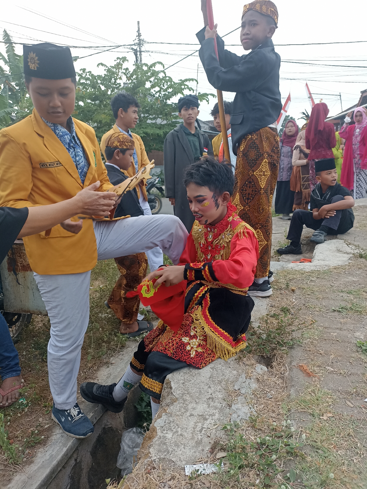 The image depicts an outdoor scene in Banyuwangi, Indonesia, likely during a cultural event or parade. In the foreground, a young boy dressed in a vibrant red and gold traditional outfit, with elaborate face paint, sits on a stone. He is holding a red cloth with green embroidery. To his left, another young person wearing a yellow jacket and a black peci (cap) is leaning forward, interacting with the seated boy. The yellow jacket has text that reads "MOCK READY ARTA." A few other children and adults are visible in the background, some in traditional attire and others in more casual clothing. Indonesian flags are present in the background, suggesting a patriotic or celebratory context. The environment appears to be a roadside with dry grass and a concrete drainage ditch in the immediate foreground. The overall impression is a snapshot of a community gathering, possibly involving a performance or procession.