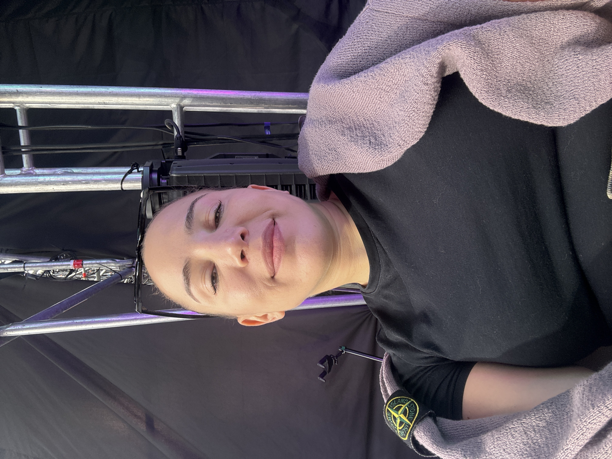 In this medium shot selfie, a young woman with her hair pulled back and light makeup is looking at the camera, her head tilted slightly. She is wearing a black t-shirt with a Stone Island logo patch on the left sleeve, and a light purple towel is draped over her left shoulder and arm. The background suggests a backstage area or event setup, with black fabric drapes and metallic scaffolding with cables and lights. The lighting is artificial and casts a slight purple hue in some areas. There are no visible landmarks or context indicating the specific location in Khabarovsk, Russia.