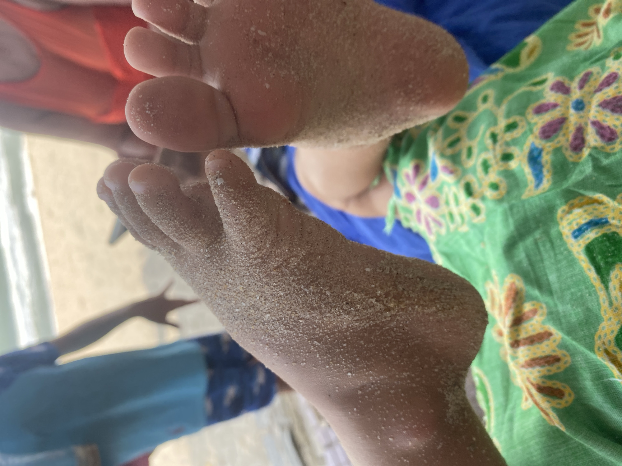 A close-up shot focuses on two sandy feet, one with the sole facing the camera, the other positioned at an angle, revealing the toes. Both feet are covered in fine, light-colored sand. In the background, out of focus, a person in a bright orange garment is visible, and further back, another person in a blue t-shirt and patterned shorts stands near the ocean. The environment appears to be a beach, suggesting the location is Mũi Né, Vietnam, known for its sandy shores. A green fabric with a floral pattern is partially visible in the foreground.