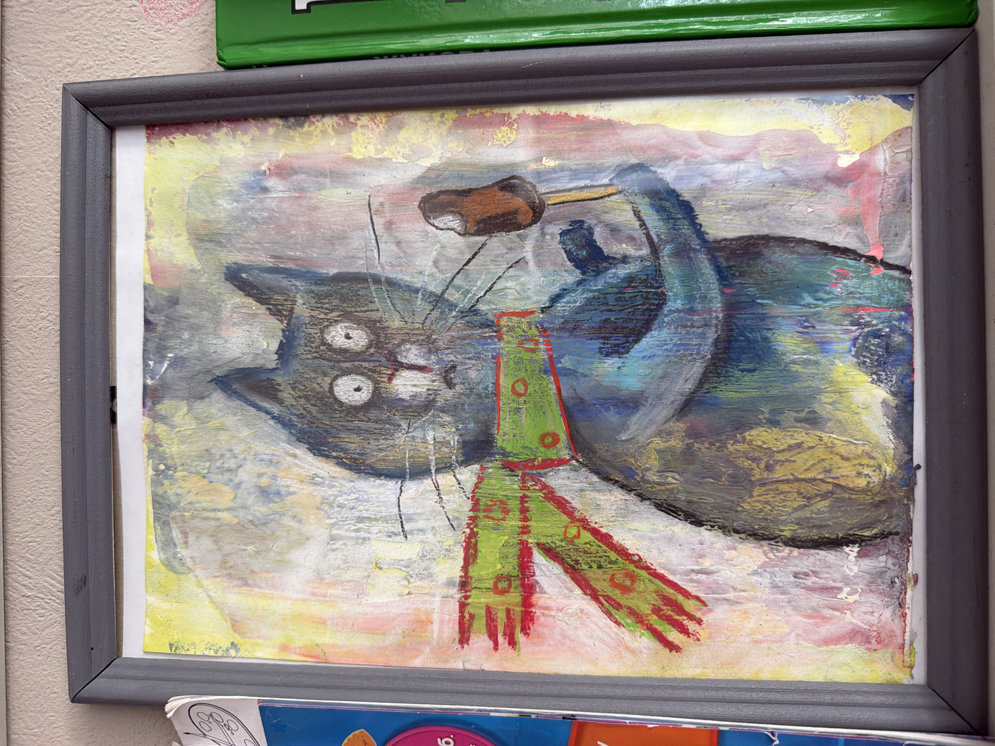 A framed, abstract painting of a cat with a green scarf and holding a popsicle is displayed on a wall. The cat is dark blue with wide, surprised eyes and black whiskers. The background is a chaotic mix of yellow, pink, white, and blue paint. A green book is visible to the right of the painting. The wall has a textured finish, and the frame is gray. There is no visible indication of the location in Ufa, Russia.
