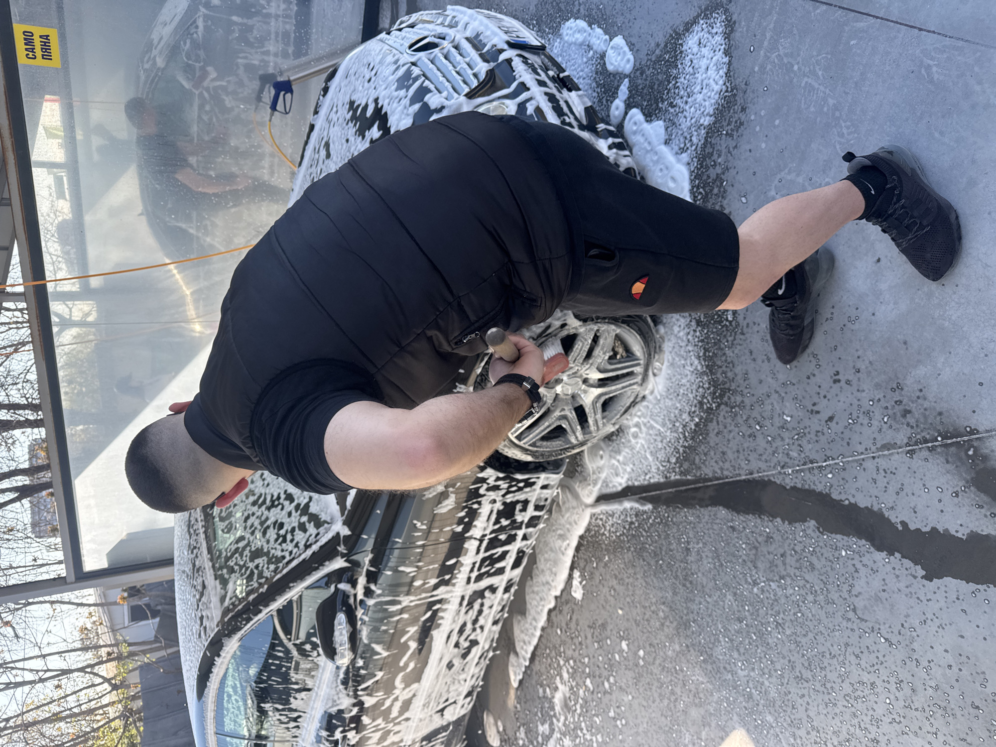 A man washes a dark-colored car at a self-service car wash. The man is wearing black shorts, a black top, and black shoes. He is using a brush to clean the car's wheels. The car is covered in white foam. There is another person in the background washing another car. The car wash is located in Plovdiv, Bulgaria, based on the Cyrillic script on a sign in the upper-left corner of the image. The ground is wet. There are trees visible outside the car wash bay.
