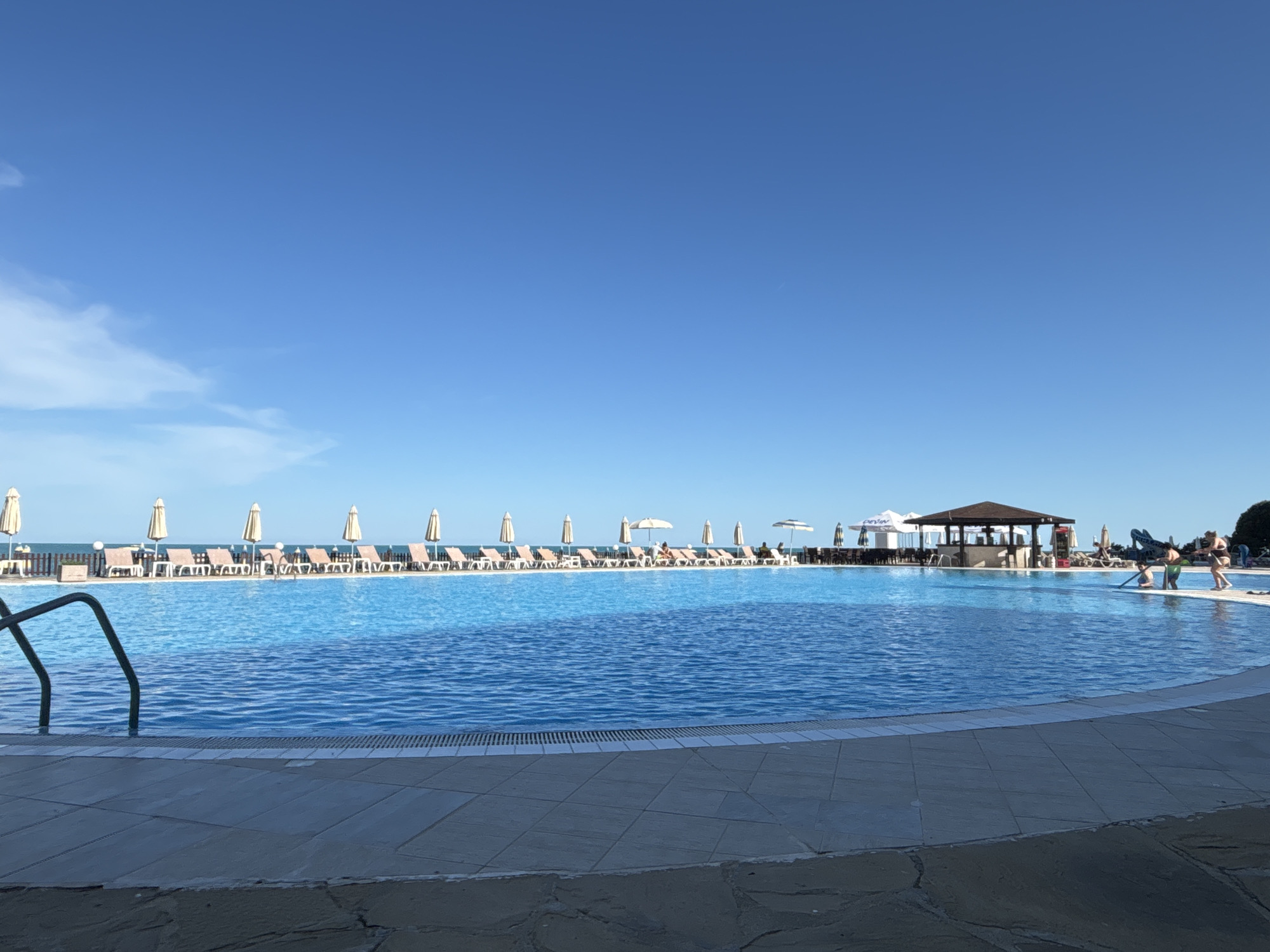 The media file depicts a large, clear blue swimming pool at a resort on a sunny day. The pool is the central focus, with lounge chairs and umbrellas lining the far edge, leading towards the ocean in the background. A covered bar area is visible behind the lounge chairs, with a few people enjoying themselves. In the foreground, metal pool ladder railings emerge from the water. The scene is set on a paved poolside area, with a view of the sea beyond the pool. The sky is a bright, clear blue, suggesting pleasant weather, likely late morning or early afternoon. Two people are visible near the far edge of the pool, one adult and one child, seemingly engaged in an activity. A portion of what appears to be a resort building with umbrellas is visible to the left of the bar. The overall impression is one of a relaxing vacation setting. No visible text is discernible.