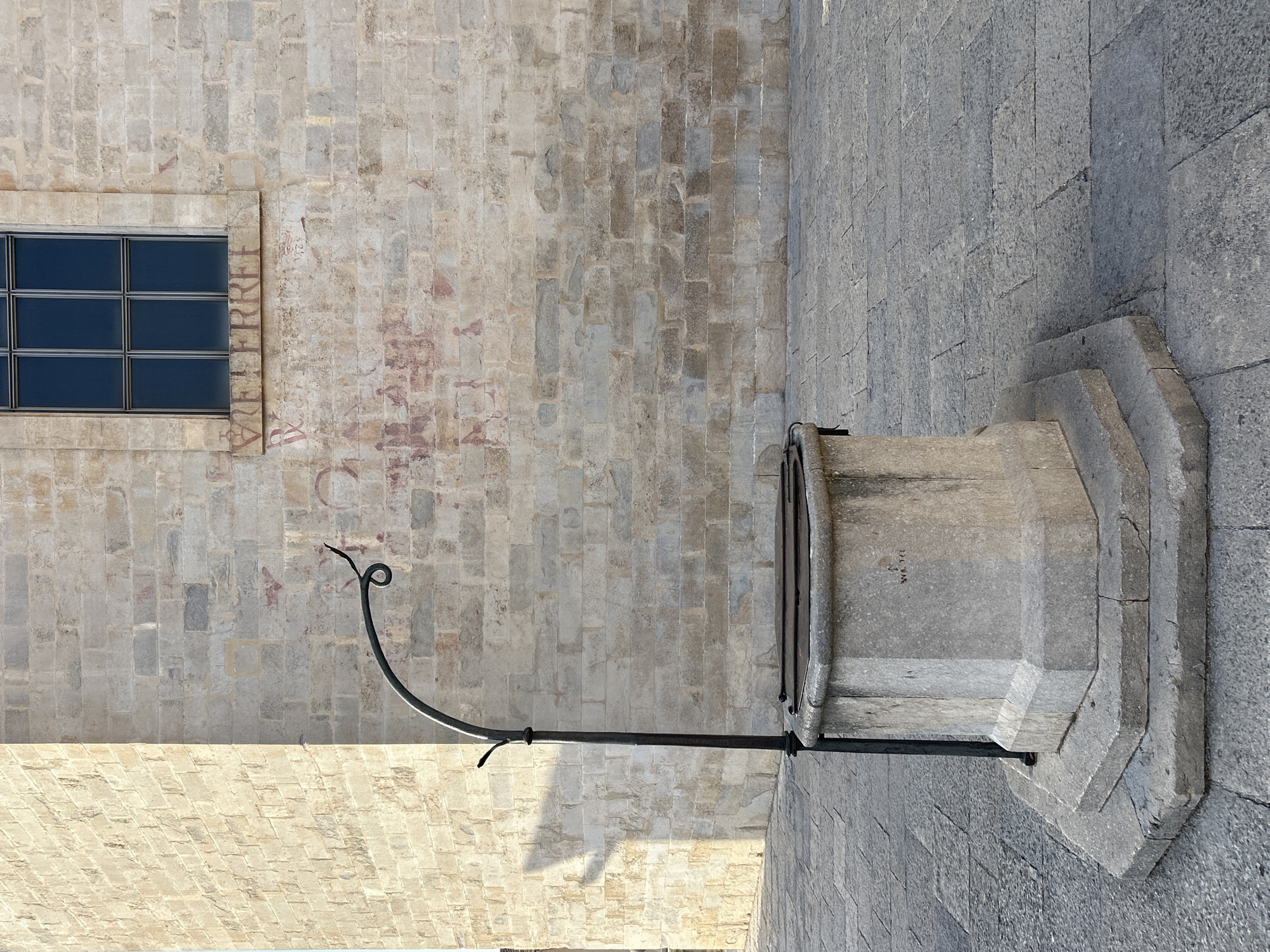 The image shows a stone well in a paved square, with a brick building in the background. The building has a window with multiple panes and some faded red markings on its wall. The well is octagonal in shape and has a metal structure extending upwards and curving over it. There is a shadow cast by a building on the left side of the image onto the well's structure and the wall behind it. The ground is paved with rectangular stones. The scene is an exterior view, likely in an urban setting. This well is located in Plaça de Sant Fèlix in Girona, Spain.
