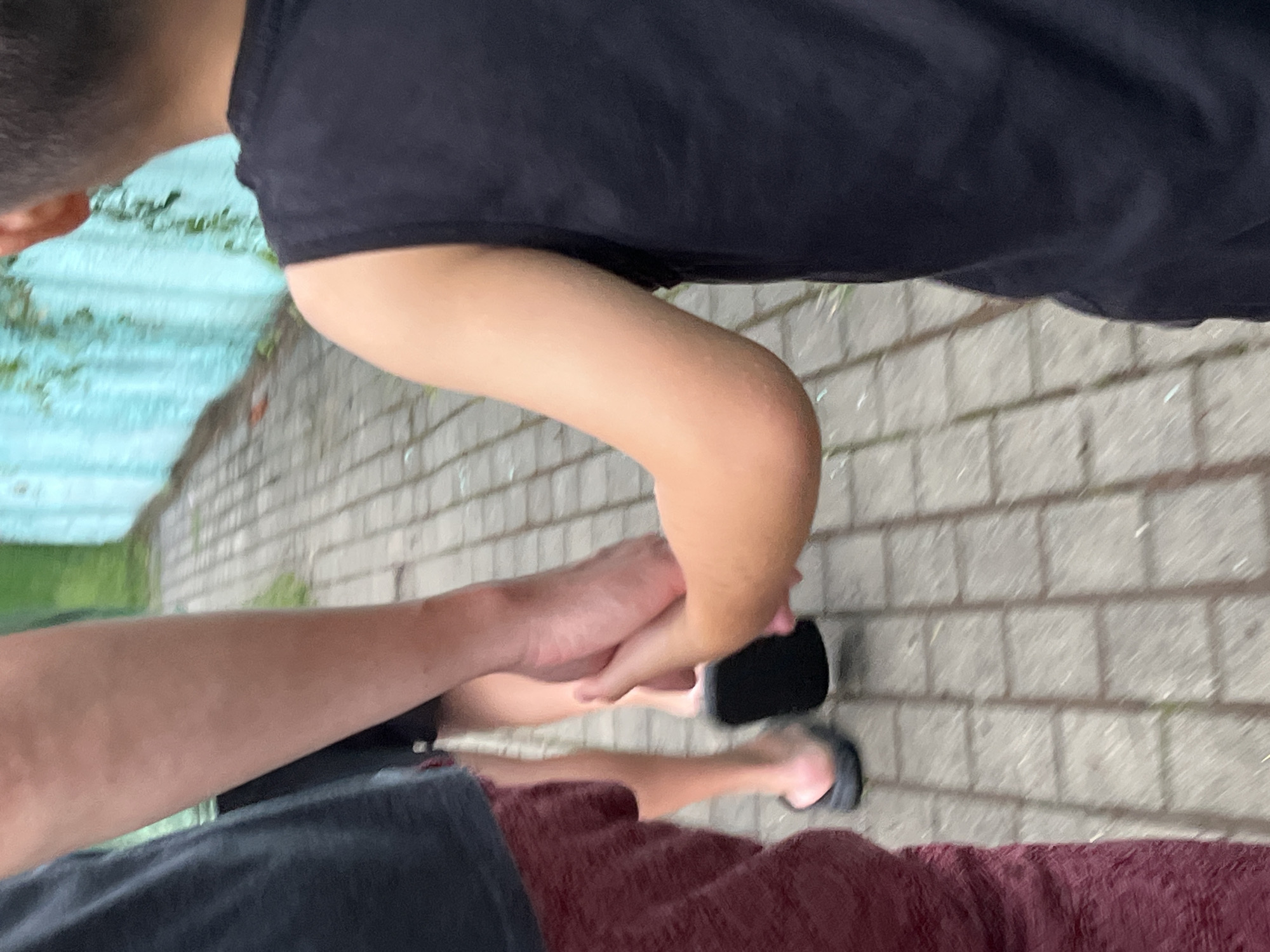 In Ureki, Georgia, a child's hand is held by an adult's hand. They are walking on a paved sidewalk beside a turquoise wall with green vines. The adult is wearing a dark t-shirt and burgundy shorts, while the child is wearing a black tank top. The child's arm is visible, and the adult's arm and hand are clasped around the child's hand. The sidewalk is made of grey square tiles. The scene suggests a peaceful moment between two individuals.