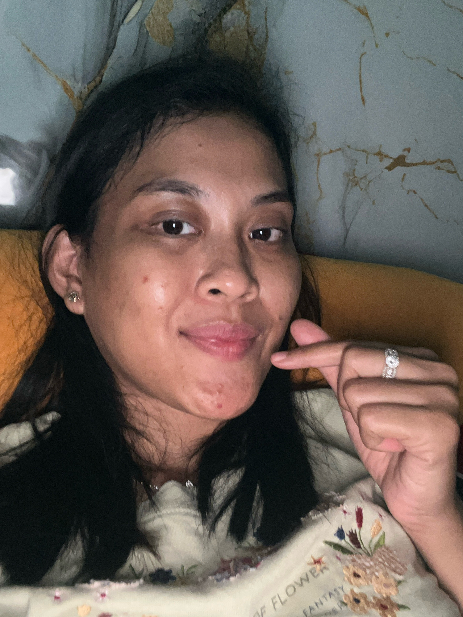 A close-up, selfie-style photograph shows a woman lying down, with her head resting on an orange pillow. She is looking towards the camera and smiling subtly. Her dark hair is down and partially covers her face. She is wearing a light-colored shirt with floral embroidery. On her left ear, she has a small, silver stud earring. Her right hand is raised, with her thumb and index finger forming a small heart shape, positioned near her chin. She is wearing two rings on her right hand: a larger, elaborate ring on her ring finger and a thinner band on her middle finger. The background appears to be a wall with a textured, marbled pattern in shades of blue and gold. The image quality is slightly grainy. The location is identified as Kecamatan Pati, Indonesia.