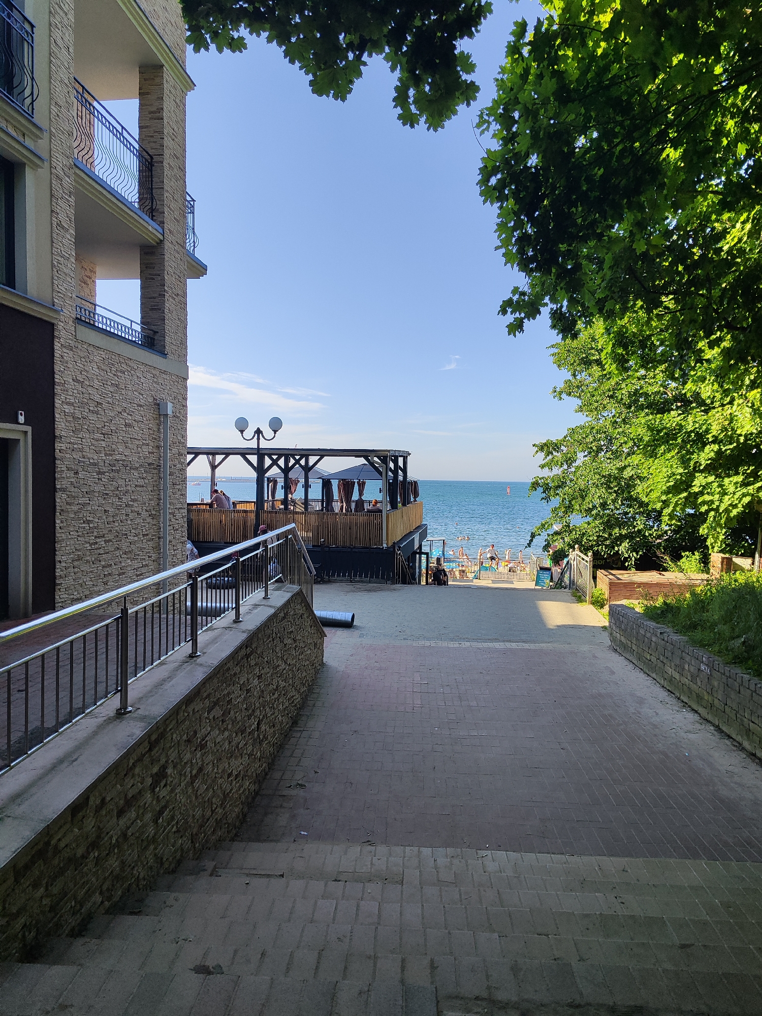 A sunny day at a beach resort in Pionersky, Russia. A paved walkway with stairs leads down towards the sea, flanked by a modern building with balconies on the left and lush green trees on the right. The building has a textured stone facade. A metal railing follows the walkway's incline.
Further down, a wooden deck area with pergolas and seating is visible, with people enjoying themselves. Beyond the deck, the blue sea stretches to the horizon under a clear sky. Several people are visible on the beach and in the water, indicating a lively atmosphere. The time of day appears to be late morning or early afternoon, given the bright sunlight casting shadows. The weather is pleasant and warm.
There is no visible text in the image. The scene evokes a sense of relaxation and vacation.