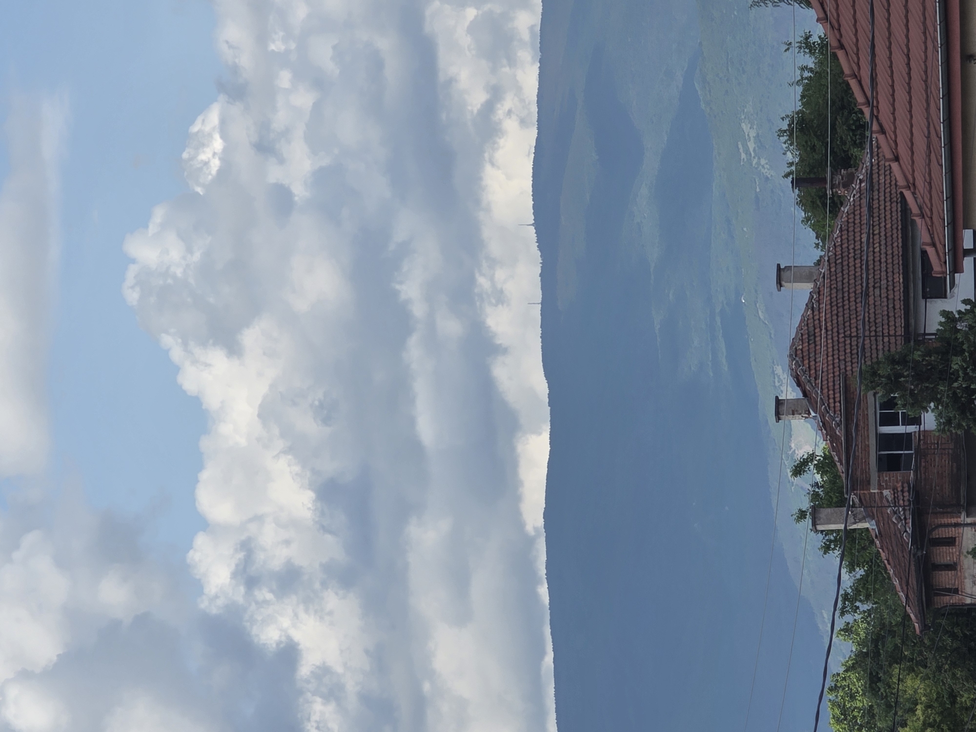 The image depicts a landscape in Yagodovo, Bulgaria, with a combination of natural and built environments. On the left, a sky with light blue patches and fluffy white clouds is visible. A dark green mountain ridge occupies the center of the frame. On the right side, the edge of a building with a red-tiled roof is seen. The building appears to be made of red brick and has a few windows. Some power lines run in front of the building. Green trees partly obscure the building's details.