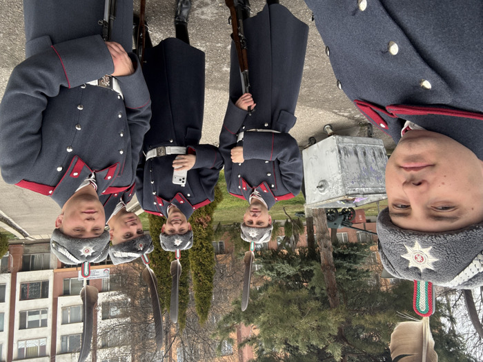 Six male individuals, appearing to be young adults, are depicted in a selfie taken on a paved pathway at the Georgi Stoykov Rakovski Military Academy in Sofia, Bulgaria. They are dressed in formal military attire: dark gray long overcoats featuring red collars, shoulder boards, and cuffs, silver-colored buttons, and white belts with silver buckles. Each wears a gray fur-brimmed hat adorned with a silver star insignia, a white braided cord, and a tall dark feather plume with a red, white, and green striped base at its base. Five of the individuals hold wooden-stocked bolt-action rifles in a vertical position. The individual positioned third from the left holds a white smartphone in his left hand. In the background, a large, weathered gray metal wheeled container is visible, along with a small vintage artillery piece. Behind them stand multi-story institutional buildings with beige facades and several bare deciduous trees under an overcast sky.