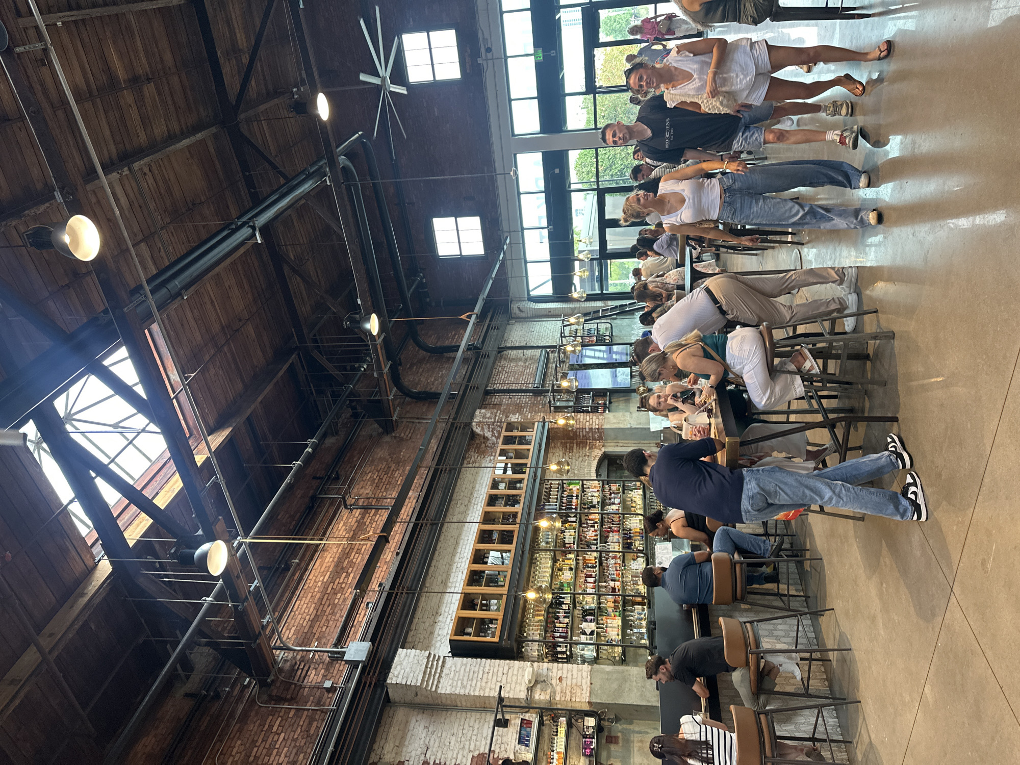 The interior of a bustling establishment, possibly a restaurant or bar, is captured with a wide-angle view. The structure features a high, wooden-beamed ceiling, exposed brick walls, and abundant natural light streaming through large windows. A prominent feature is a well-stocked bar with shelves filled with bottles. Patrons are seated at tables and the bar, engaged in conversation and dining. The ambiance is lively and social. The location is in the city of Tampa, United States of America.