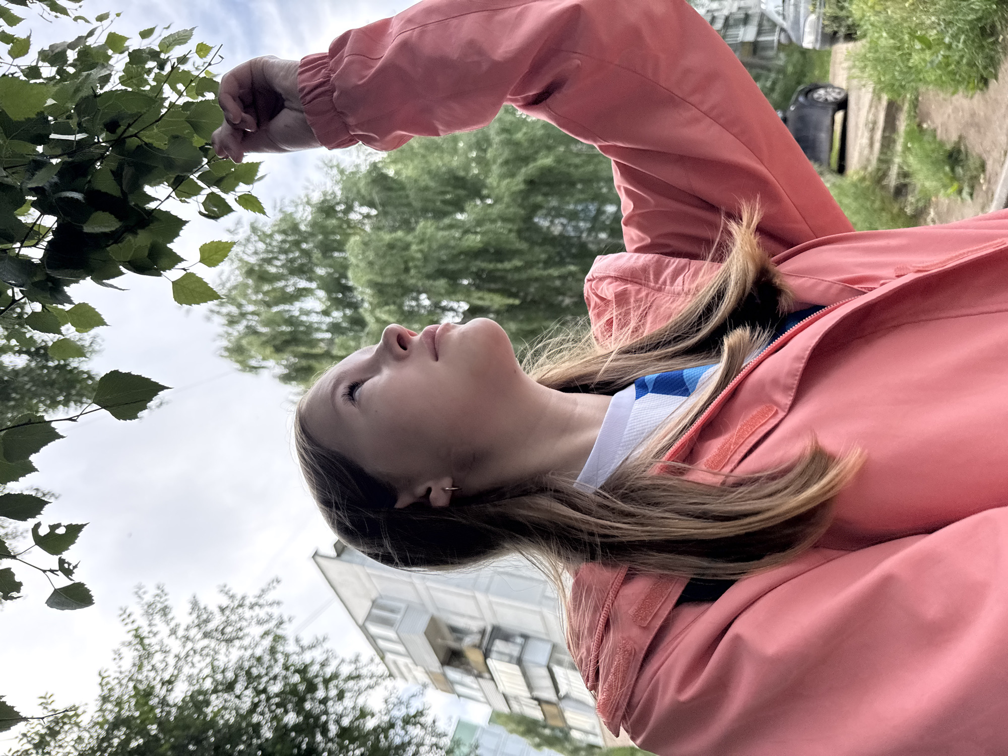 A young female with long, light brown hair, wearing a coral-colored jacket over a white shirt and blue tie, looks up at tree leaves outdoors. She is in an urban environment, with a grey, multi-story apartment building visible in the background. Two cars are parked on a dirt path, with green grass and foliage surrounding the area. The sky is overcast. The image appears to be taken on a street or pathway in the city of Chaikovsky, Russia.