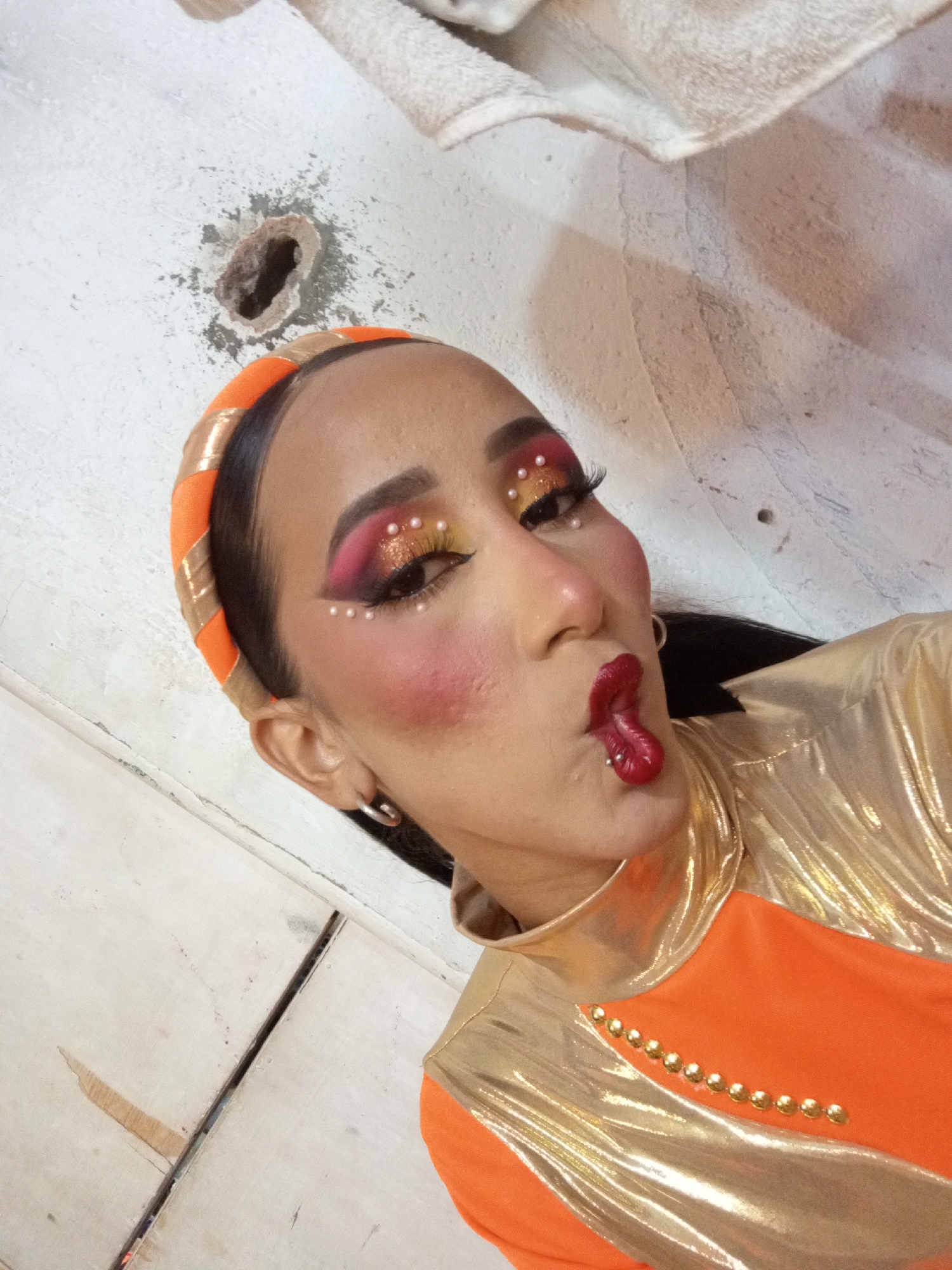 A woman with heavy makeup (pink and orange eyeshadow, false eyelashes, rouge, pearls on the eyelid and a lip piercing) wears an orange headband and a gold and orange outfit. She is pursing her lips toward the camera. The background is a white wall with a hole and a white towel. Based on previous exchanges, this is likely in Porlamar, Venezuela.
