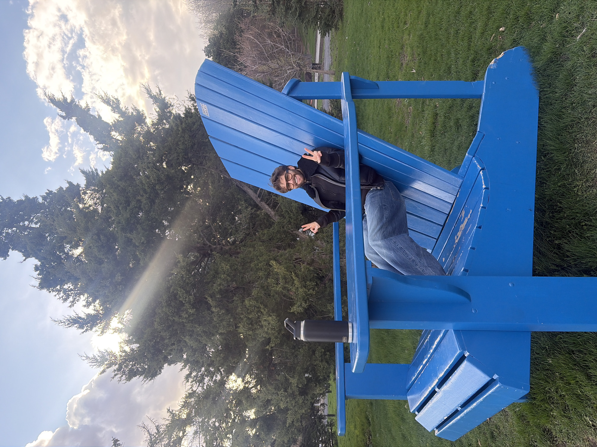 A man sits in a large blue Adirondack chair outdoors. He wears glasses, a black jacket, and jeans. He holds up two peace signs with both hands. A black water bottle rests in the chair's armrest cup holder. The chair sits on a grassy area. Trees are in the background, and the sky has clouds and sun. The location is not discernible from the image.
