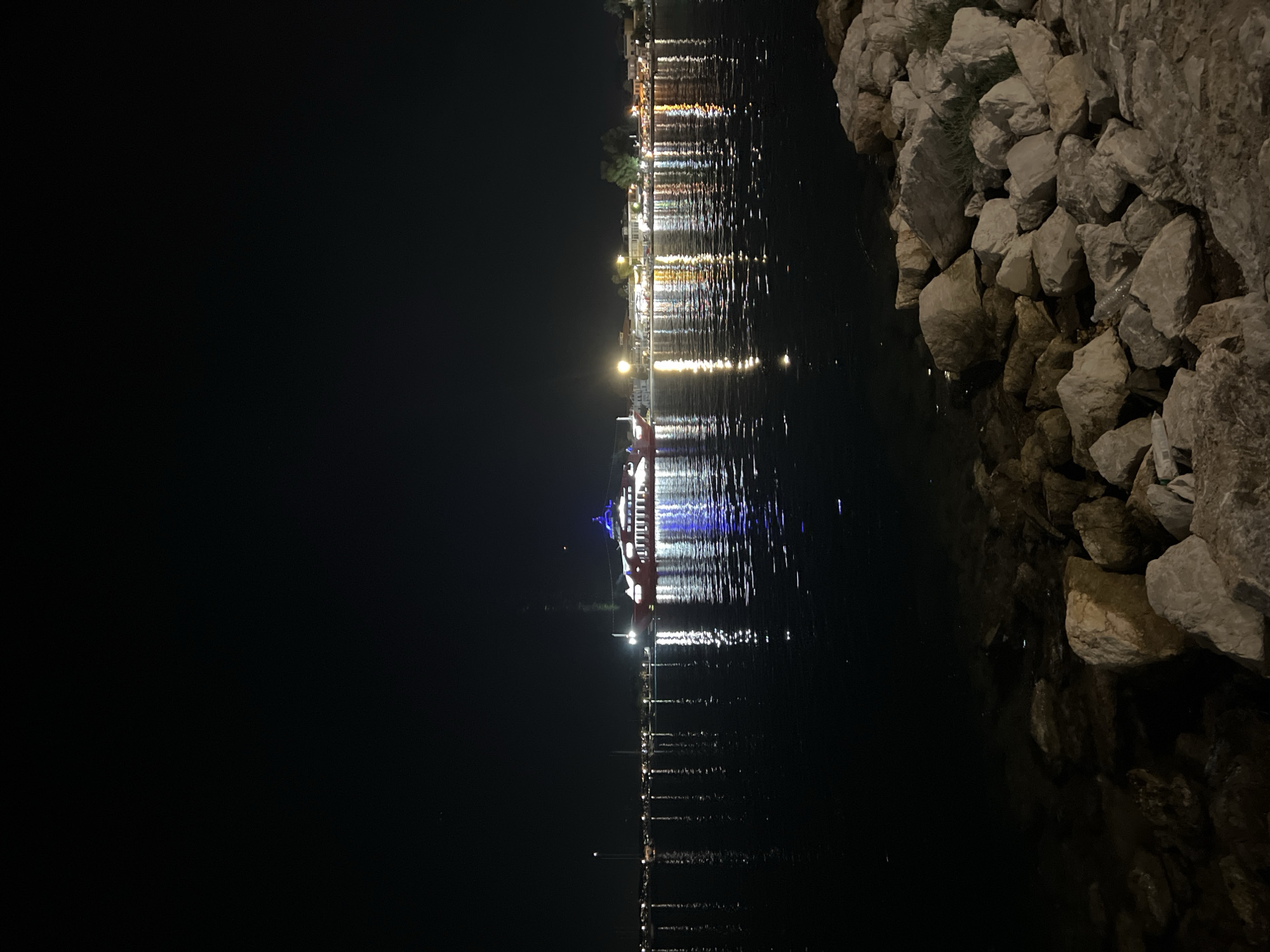 A night scene depicts a waterfront in the city of Eretria, Greece. The foreground features a rocky shoreline with large, light-colored stones. Beyond the rocks, a calm body of water reflects the lights from a town and a large ferry illuminated with blue and white lights. The ferry is positioned on the right side of the water, with its lights creating a strong reflection. In the distance, along the waterfront, buildings are visible with numerous lights, suggesting a populated area. The sky above is dark and clear. The overall impression is of a peaceful, illuminated evening by the sea.