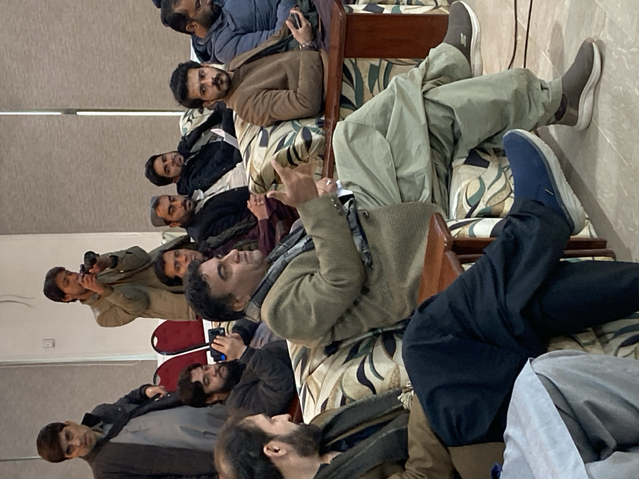An indoor scene in Quetta City Tehsil, Pakistan, depicts a gathering of approximately ten adult males. Most individuals are seated on patterned sofas featuring cream, brown, and dark blue designs with wooden armrests. The walls are light gray/beige, and large windows with light brown vertical blinds are visible in the background. The floor is a light-colored tile or carpet.
In the central foreground, a middle-aged man with dark hair and a light brown/olive blazer, a patterned gray scarf, and light green shalwar is seated, gesturing with his raised right hand, seemingly conversing. He wears brown slip-on sneakers. To his immediate left, a man with a dark beard is partially visible, wearing a dark brown garment. To his immediate right, a young man with a mustache and short beard, wearing a brown jacket, is seated, holding a smartphone in his left hand. He wears gray slip-on sneakers.
Further back, on the left, a man with a dark beard is seated, holding a smartphone horizontally. To his right, another man is partially visible. Next to him, a man with a dark beard wears a light cap and a dark jacket. To his right, a man with a dark beard is seated, holding a white sheet of paper. Another man is seated further right.
In the background, a man in a dark jacket stands on the left. To his right, another man in a light brown jacket stands, holding a black DSLR camera with both hands, aiming it towards the right side of the frame. Two red chairs with white frames are partially visible in the left background.
