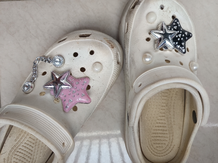 The image displays two white, well-worn, rubberized clogs, consistent in style with the Crocs brand, resting on a light-colored, possibly ceramic or porcelain, tiled floor. The scene is located within an unspecified indoor or sheltered outdoor environment in Gotha, Germany. The clog on the left is adorned with three decorative elements: a silver-toned chain charm featuring two clear, faceted faux diamonds, and a layered star charm composed of a larger pink star with silver dot patterns, topped by a smaller, reflective silver star. The clog on the right is embellished with two small white, pearl-like bead charms and a layered star charm consisting of a larger black star with white dot patterns, overlaid by a smaller, reflective silver star. Both clogs exhibit visible dirt, scuff marks, and wear, particularly on their surfaces and inside footbeds, which display textured patterns. No individuals are present, and no actions or interactions are depicted. The lighting is diffuse, casting minimal shadows.