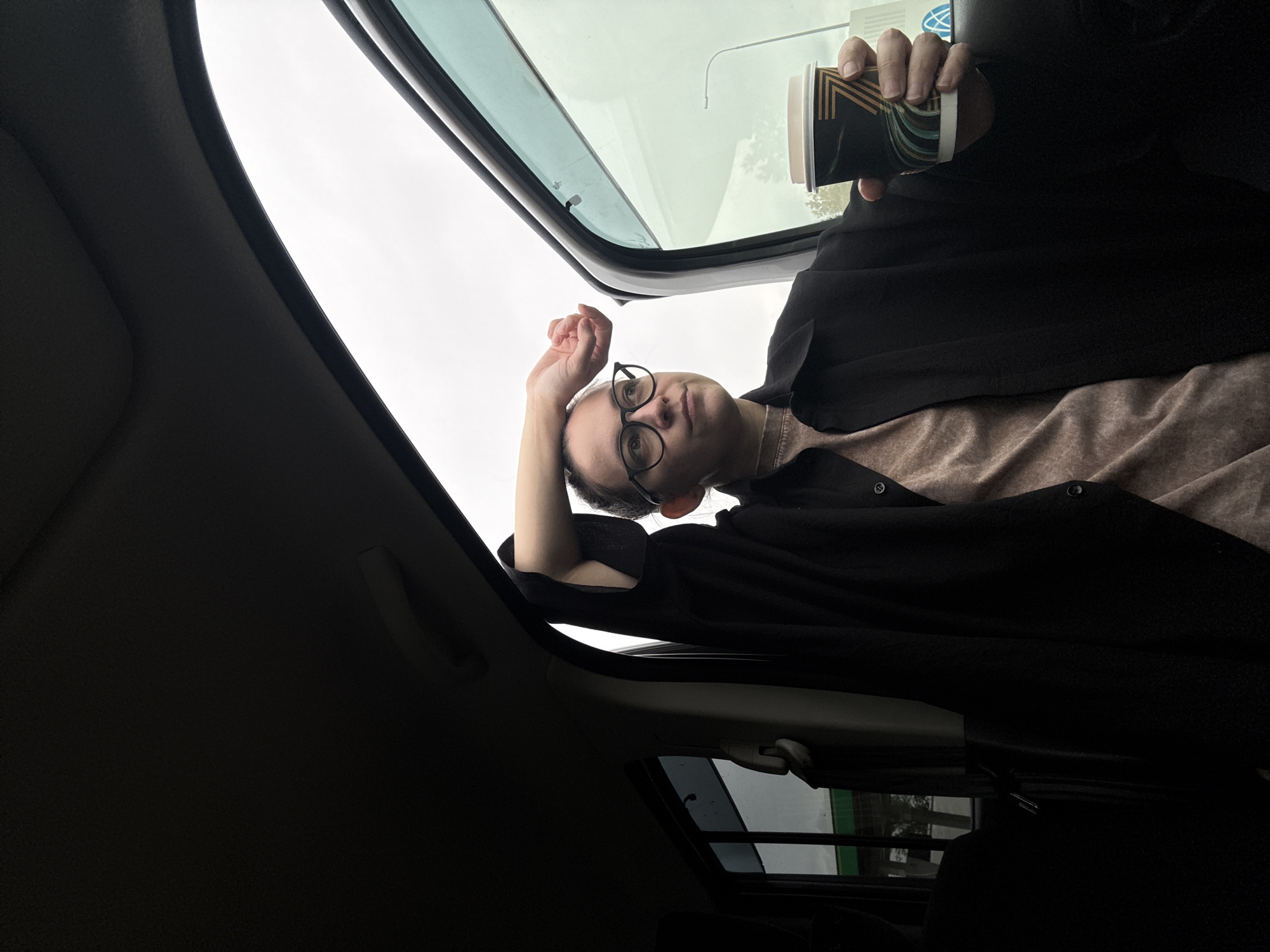 A person with short, dark hair and glasses is seen from a low angle, lying in a vehicle, possibly a car, looking up through an open sunroof. The person is wearing a light-colored shirt and a dark cardigan. They are holding a paper coffee cup in their right hand. The sky is visible through the sunroof. The interior of the car is dark, with a black ceiling and side panels. The location is not specified in the image and, based solely on the photograph, cannot be identified as Munaika, Russia.