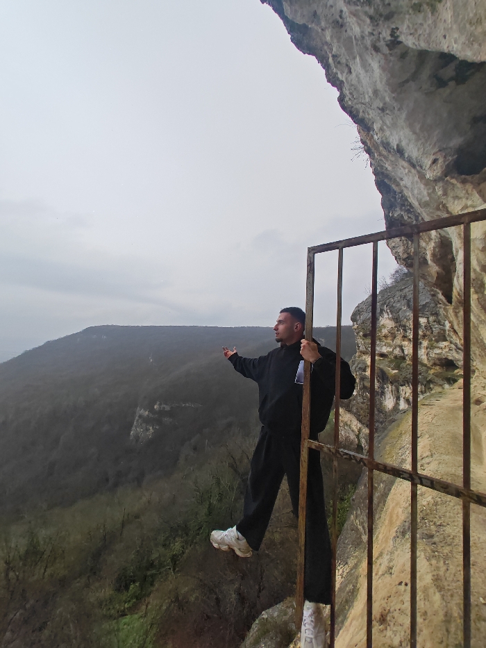 The image depicts an adult male posing on a narrow rock ledge behind a rusty metal gate, overlooking a vast forested valley on the Shumen Plateau, Shumen, Bulgaria.
The scene is set outdoors under an overcast, light grey sky. To the immediate right, a rugged, light-colored rock cliff face ascends vertically, forming a prominent overhang in the upper right corner of the frame. A weathered, tall metal gate, composed of vertical and horizontal bars exhibiting visible rust, stands in the mid-right.
An adult male, estimated to be between 20 and 35 years old, with short dark hair and a faint goatee, is positioned on the rock ledge directly behind the metal gate. He is dressed in a black hooded sweatshirt, black loose-fitting trousers, and white athletic shoes.
His left hand firmly grips a vertical bar of the metal gate. His right arm is extended horizontally to his right, with his palm open and fingers splayed, gesturing towards the expansive landscape. His left foot is elevated and rests on the lowest horizontal bar of the gate, while his right foot remains on the rocky ledge behind the gate. The male’s head is turned to his right, aligning his gaze with his extended arm and the distant valley. The background features a dense, dark green to brown deciduous forest covering extensive rolling hills and valleys, indicative of autumn or early spring foliage.