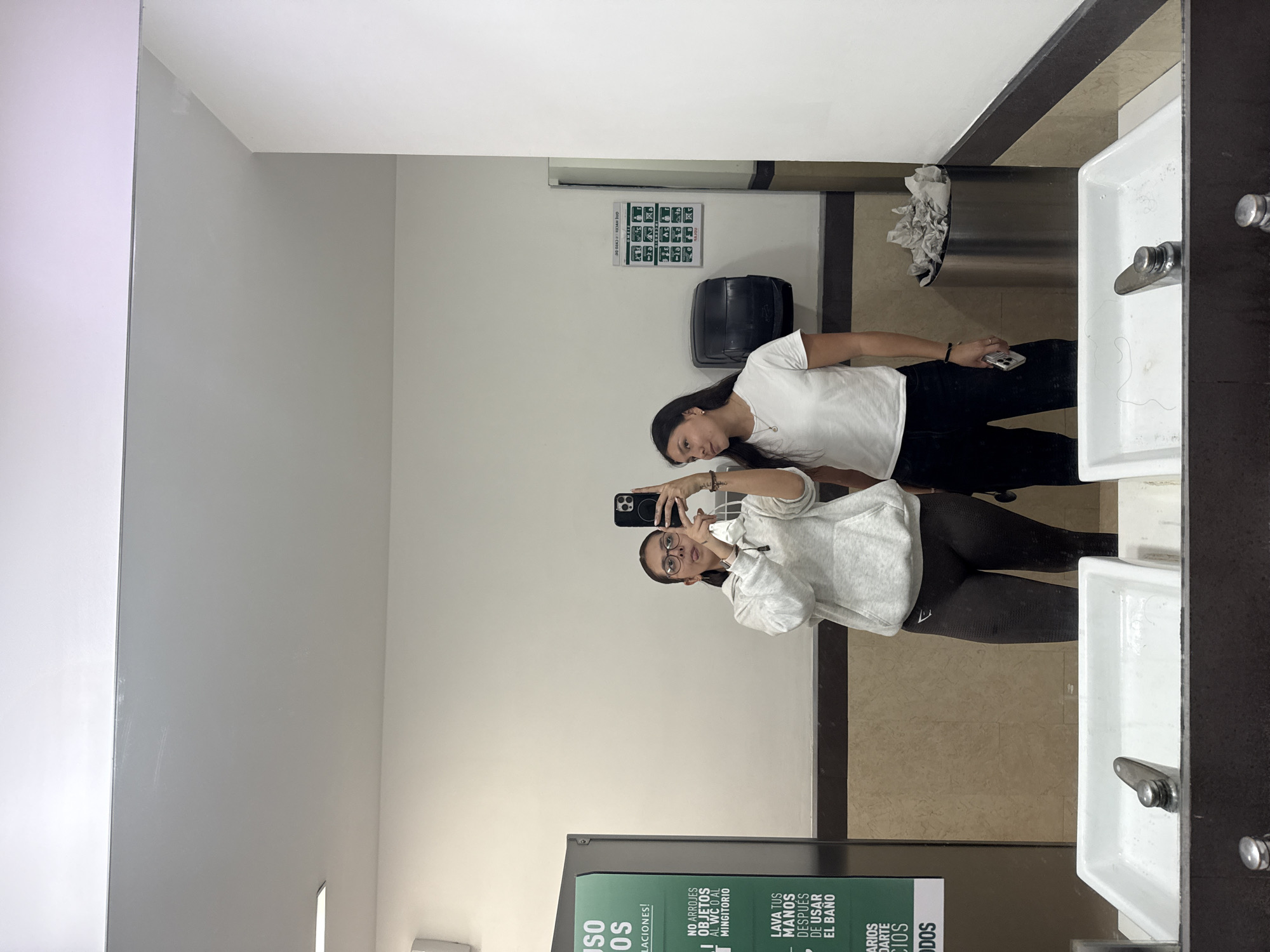 The image, rotated 90 degrees clockwise, captures two young women reflected in a large mirror inside what appears to be a public restroom. The setting features light-colored walls and a tiled floor.
On the left of the reflection, one woman is actively taking a mirror selfie using her smartphone, which has a black case. She is wearing eyeglasses, a light grey hooded sweatshirt, and dark leggings. Her dark hair is styled back. Beside her, on the right, stands another woman with long dark hair, dressed in a white t-shirt and black pants, looking towards the phone with a neutral expression.
Behind the women in the reflection, standard bathroom fixtures are visible. Two white sinks with chrome faucets are on the right, set into a dark countertop. A metallic paper towel dispenser, partially filled with crumpled paper, and a black trash can are mounted on the wall.
Several signs are present on the walls. One white sign with green pictograms and text is visible, likely outlining health or safety protocols, possibly related to COVID-19. Below this, another sign with Spanish instructions is partially visible, stating: "NO ARROJES OBJETOS AL WC O AL MINGITORIO" (Do not throw objects in the toilet or urinal) and "LAVA TUS MANOS DESPUÉS DE USAR EL BAÑO" (Wash your hands after using the bathroom). Other partial phrases like "SO OS LACIONES!" and "ARIOS DARTE CIOS ODOS" are also discernible.
The scene depicts a casual moment, typical of a public restroom environment, possibly located in San Andrés Cholula, Mexico.