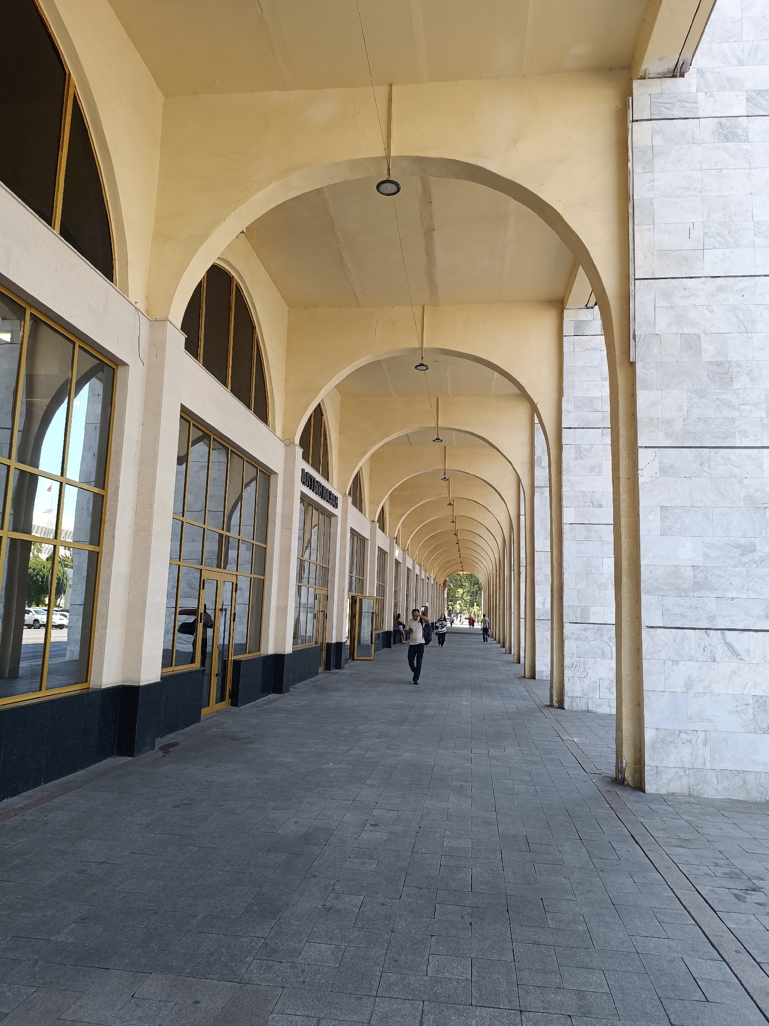 A long, arched colonnade stretches into the distance along a paved walkway in Bishkek, Kyrgyzstan. The architecture features a series of yellow-painted arches supported by columns. To the left, large windows with golden frames line the building, reflecting the sky and surrounding trees. Some of these windows have doors. To the right, the building's facade is clad in light-colored marble tiles. Several individuals are walking along the colonnade. One person in the foreground is walking towards the viewer, while others are further down the path, appearing to walk away or stand. The ground is paved with grey square tiles. The overall impression is one of a public space with a distinctive architectural style.