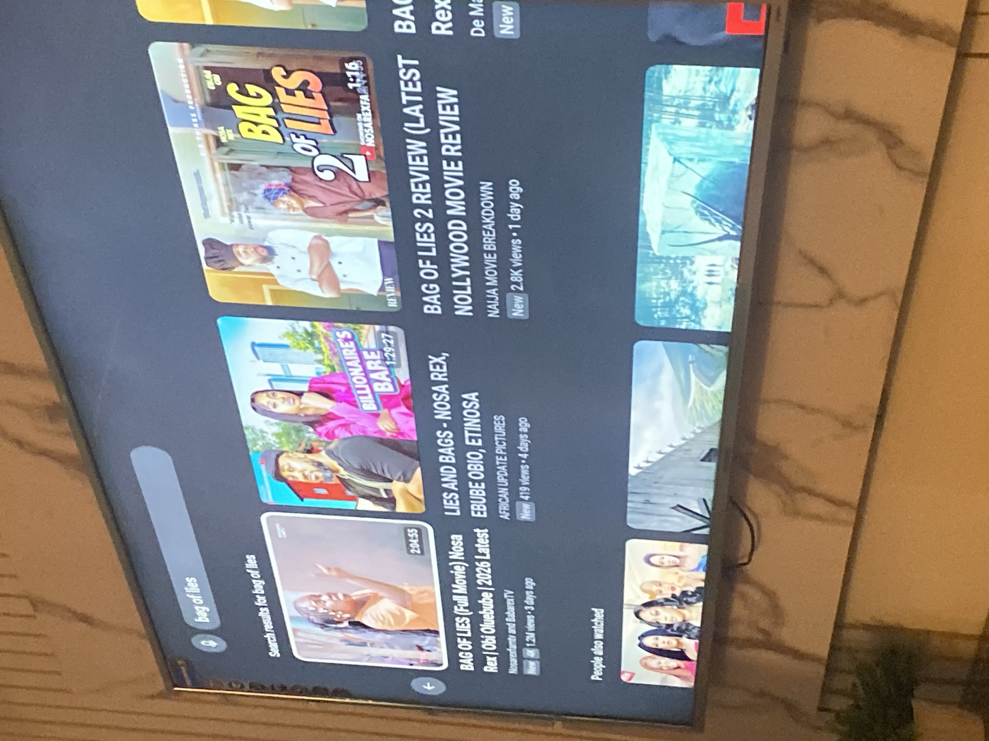 The image shows a television screen displaying search results on YouTube for "bag of lies." The screen displays several movie thumbnails and titles, including "BAG OF LIES (Full Movie) Nosa Rex" and "BAG OF LIES 2 REVIEW (LATEST NOLLYWOOD MOVIE REVIEW." There is also a list of "People also watched" and another movie titled "LIES AND BAGS - NOSA REX, EBUBE OBIO, ETINOSA." The location is not determinable from the image.