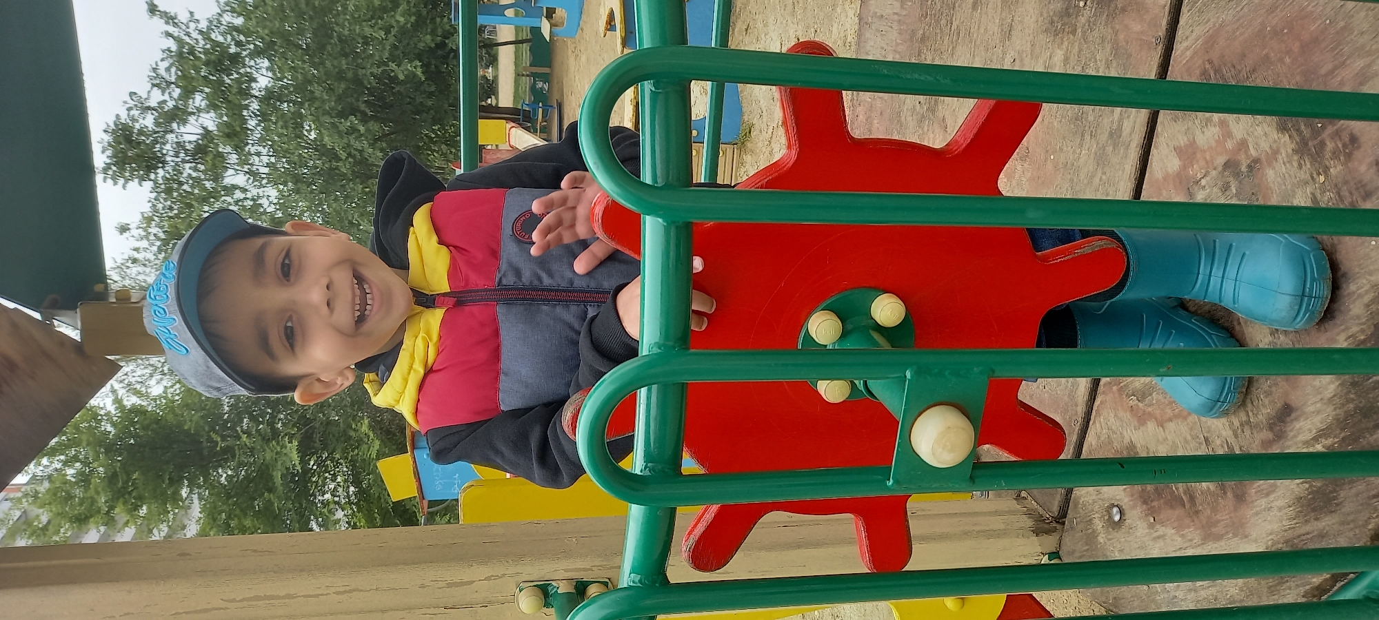 A child with a grey baseball cap and a multi-colored jacket is on a playground in Krasnodar, Russia. The child is smiling and interacting with a red and green piece of playground equipment, while wearing blue boots. Other playground equipment and trees are visible in the background.